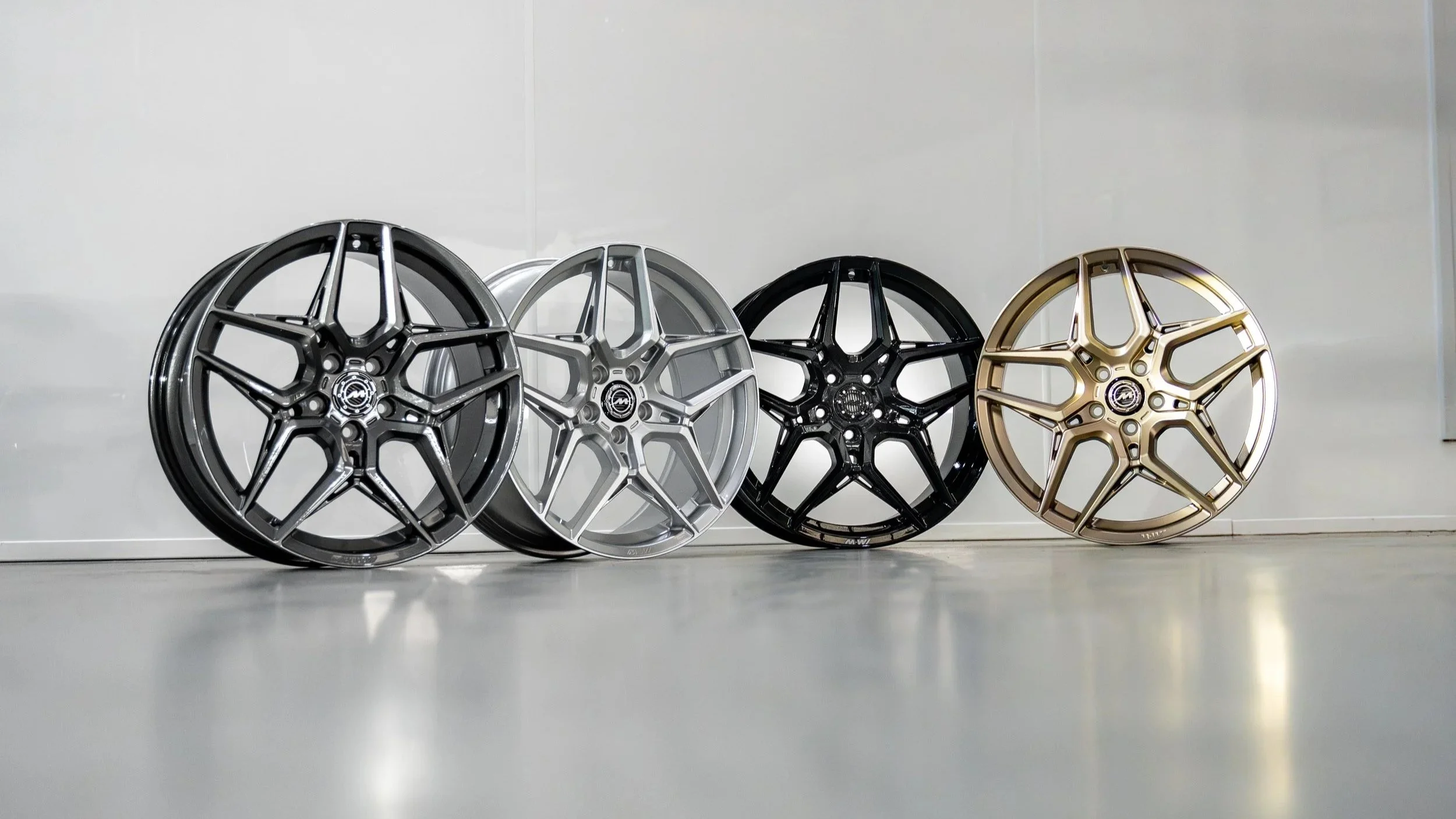 Four alloy wheels in black, silver, black, and gold finish leaning against a wall on a light-colored floor.