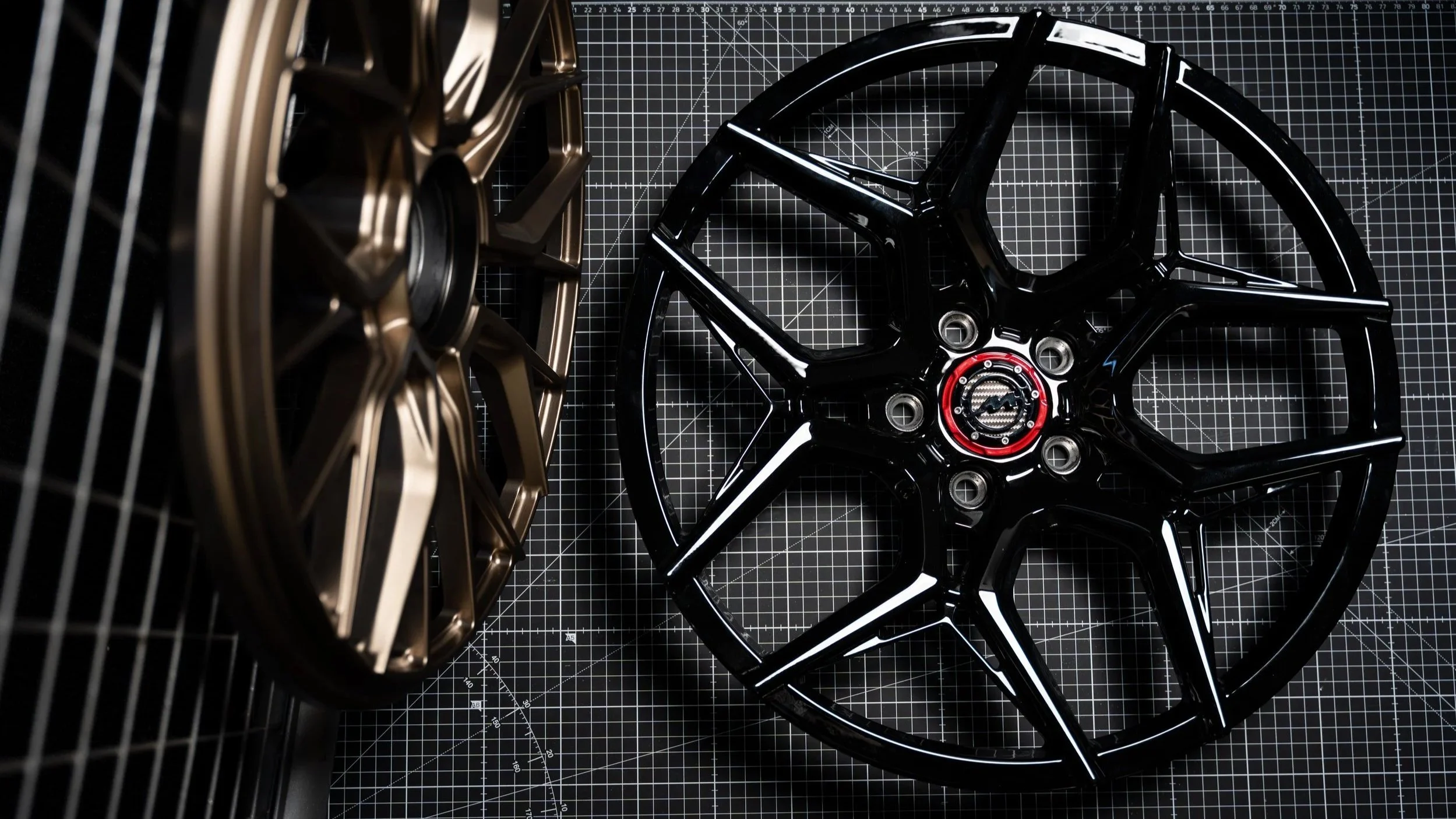 Two car wheel rims, one black and one bronze, placed on a black work surface with a grid pattern.