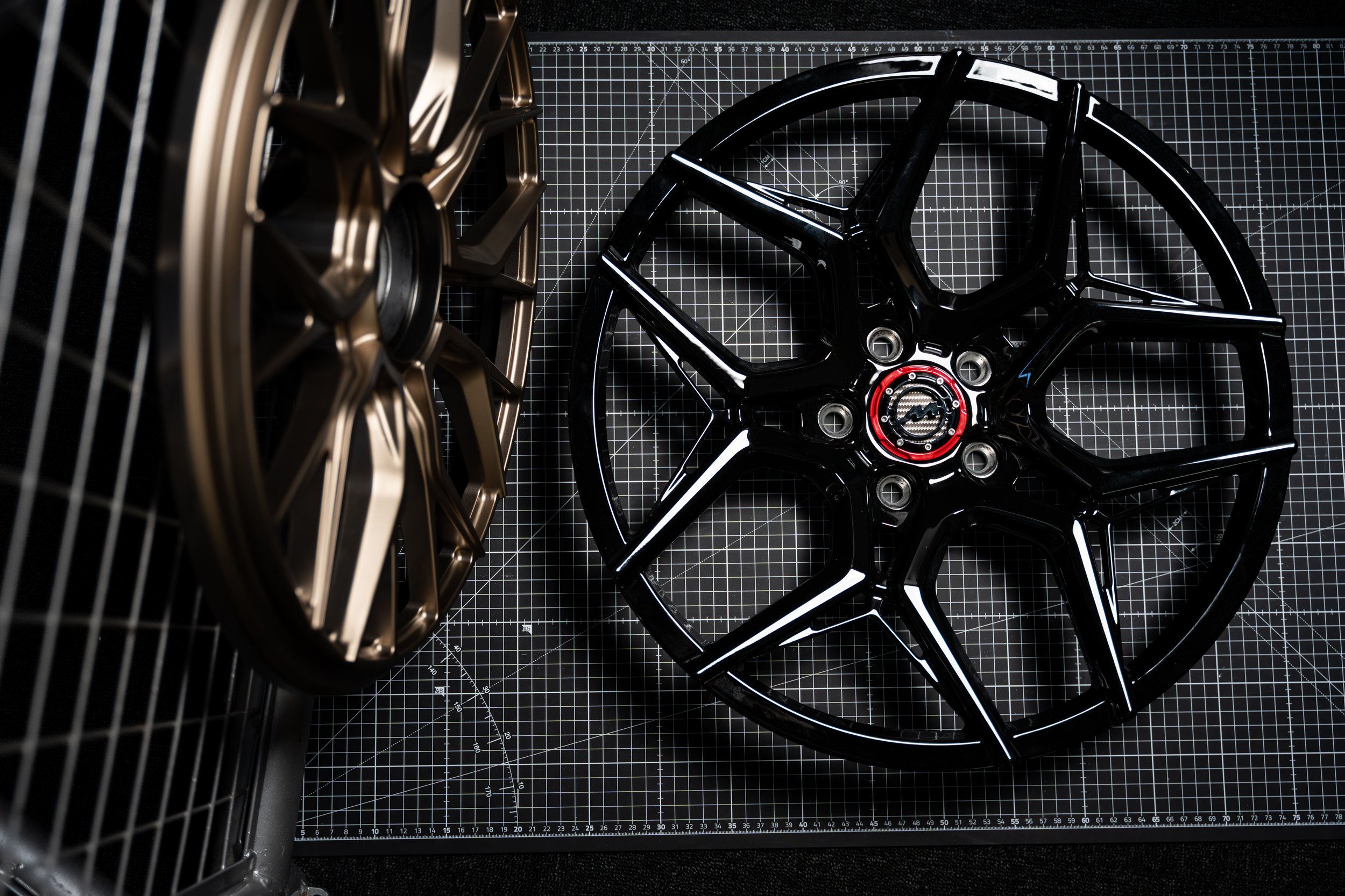 Two car wheels, one black with red accents and one bronze, placed on a black grid cutting mat with a metal grid in the background.