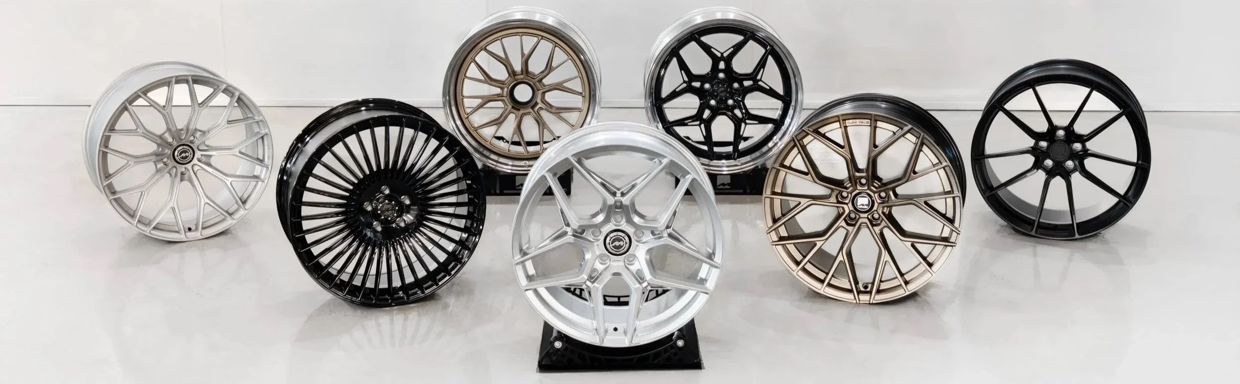 A display of nine car wheels in various designs and finishes, arranged on a white surface with a plain white background.