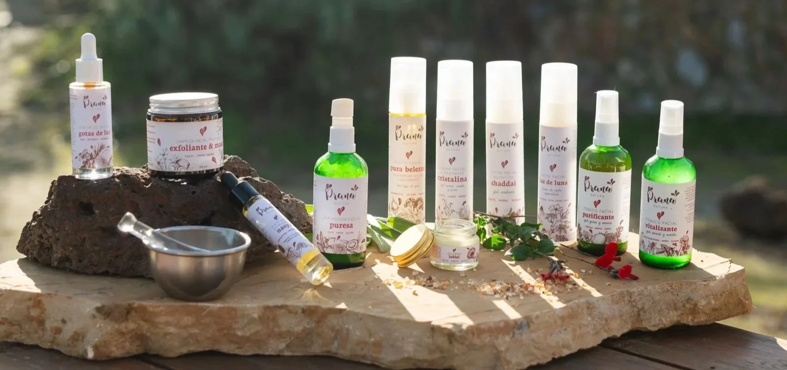 Diversos productos de belleza y cuidado de la piel en frascos y botellas de color verde, algunos con atomizador y otros con gotero, colocados sobre una superficie de piedra junto a un pequeño recipiente de metal y un frasco abierto. Fondo natural con árboles y luz natural.