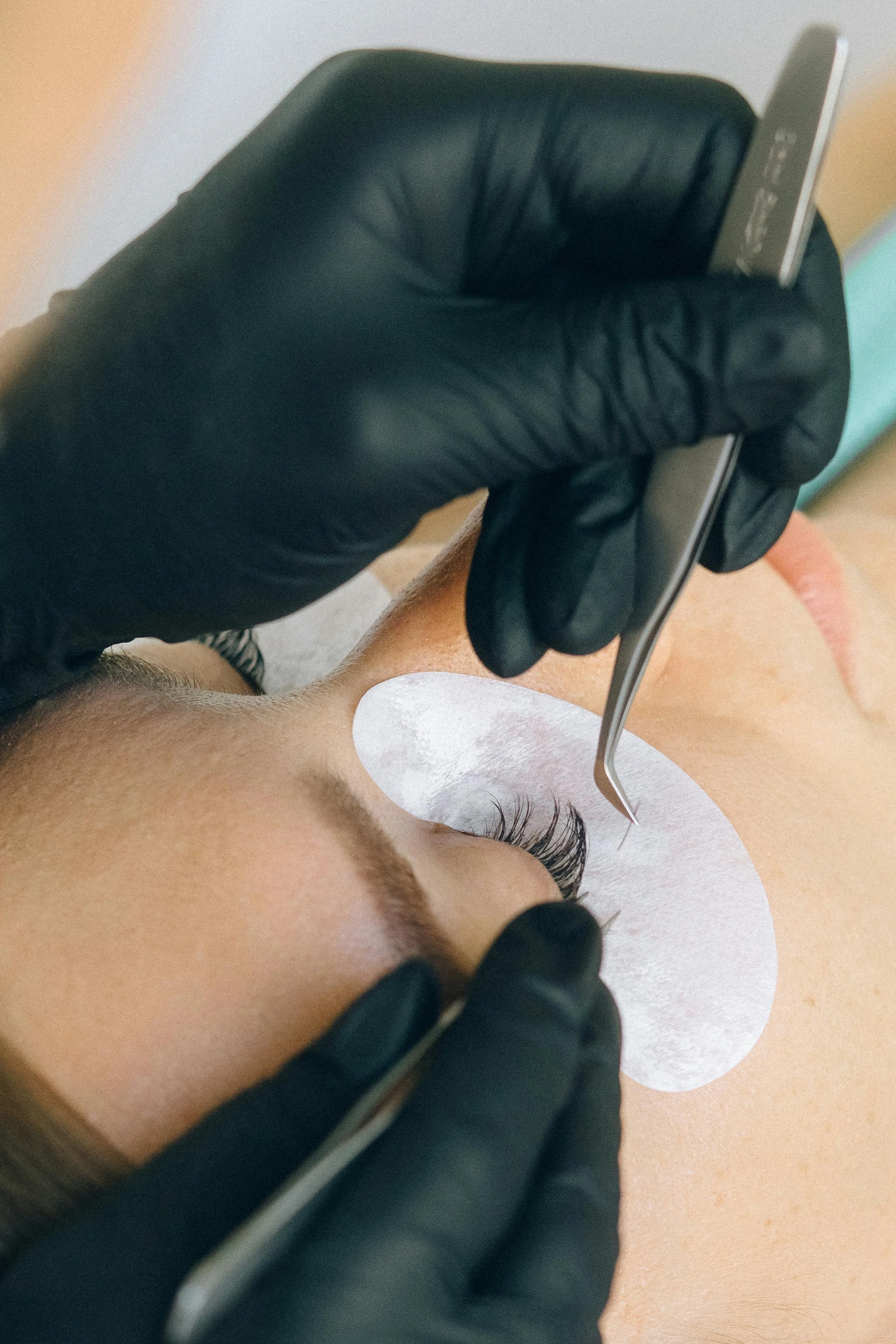 Close-up of a person receiving eyelash extension application, with a technician wearing black gloves using tweezers to attach synthetic eyelashes to a client's natural lashes, under eye protection pad on the client's eyelid.