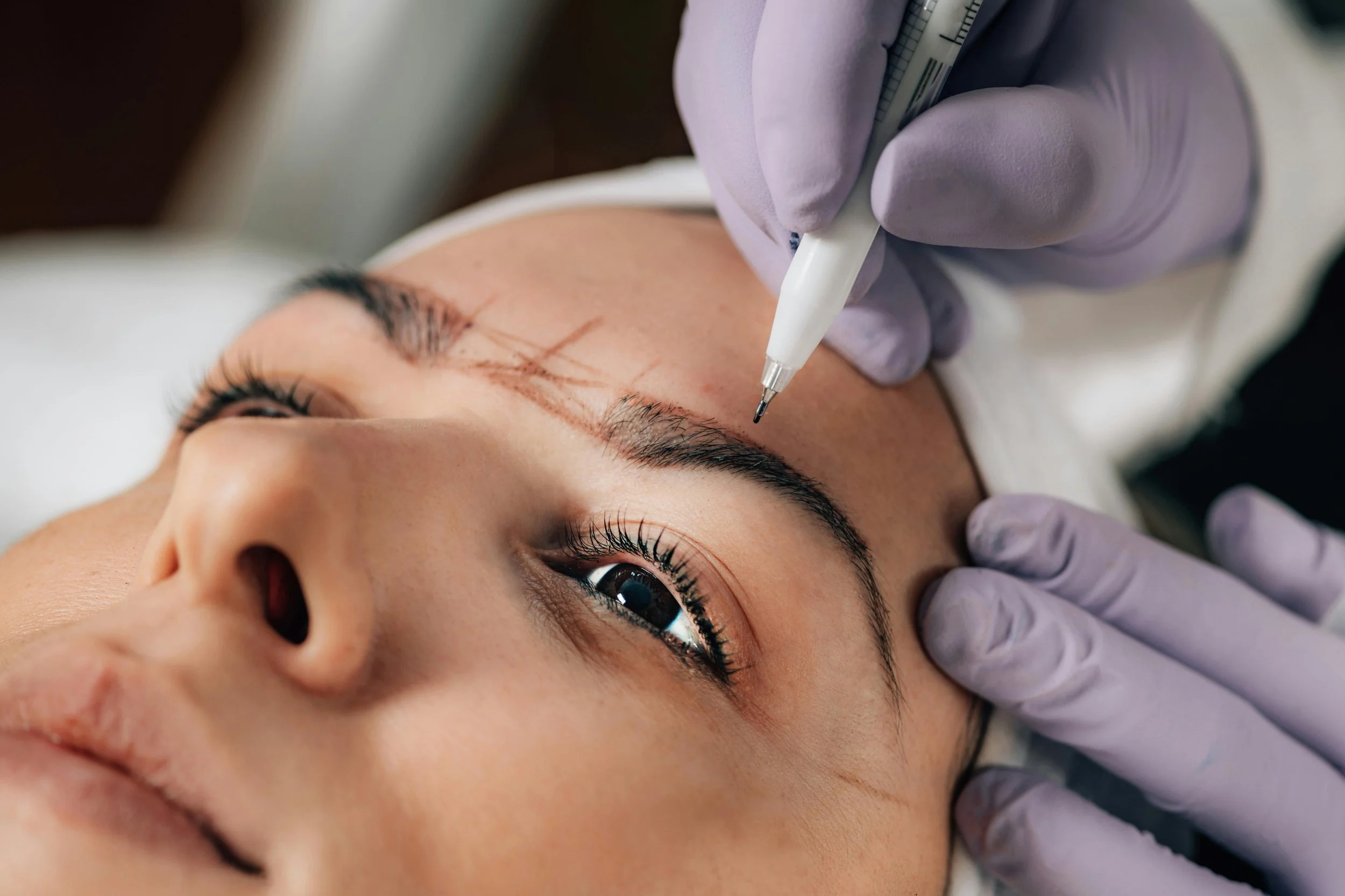 A woman receives an eyelash extension procedure, with visible markings on her eyebrow for the treatment.