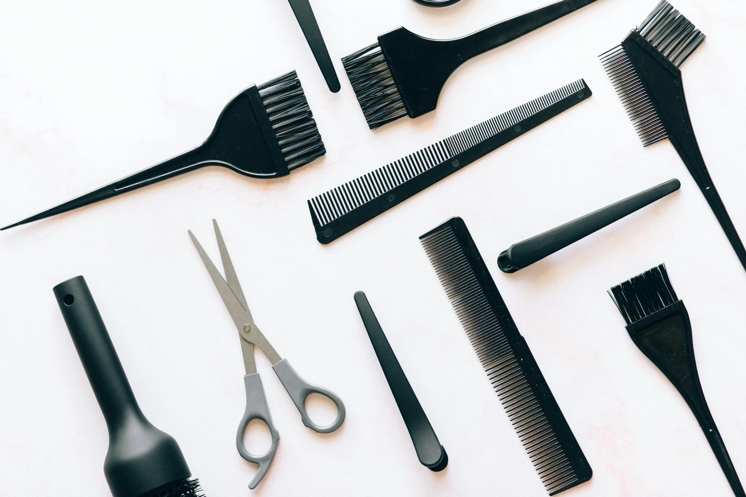 Collection of black and gray hair styling tools including brushes, combs, scissors, a hairdryer, a spray bottle, and a razor, arranged on a white background.