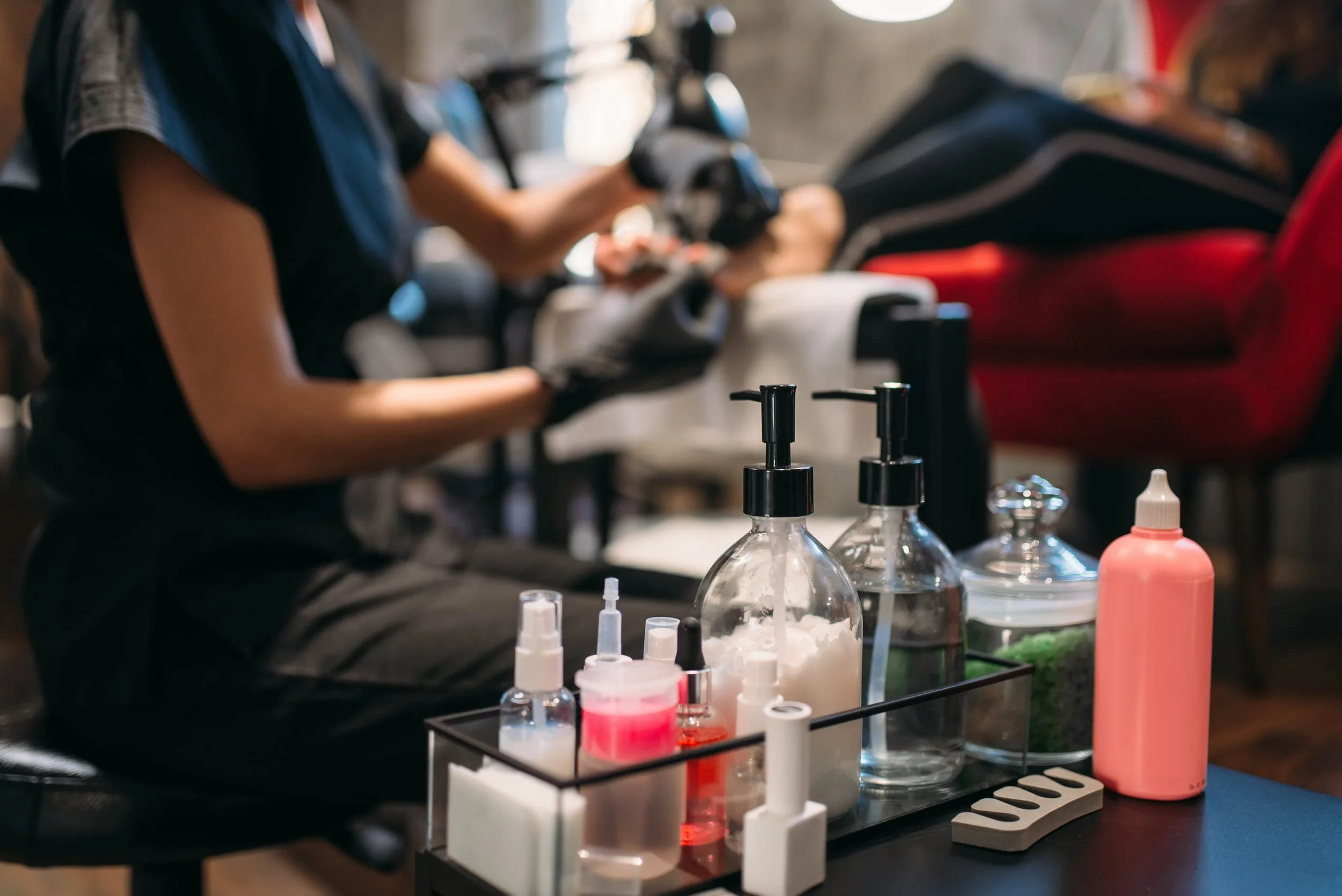 A nail technician preparing nail tools and supplies on a tray, with clients getting manicures in the background.
