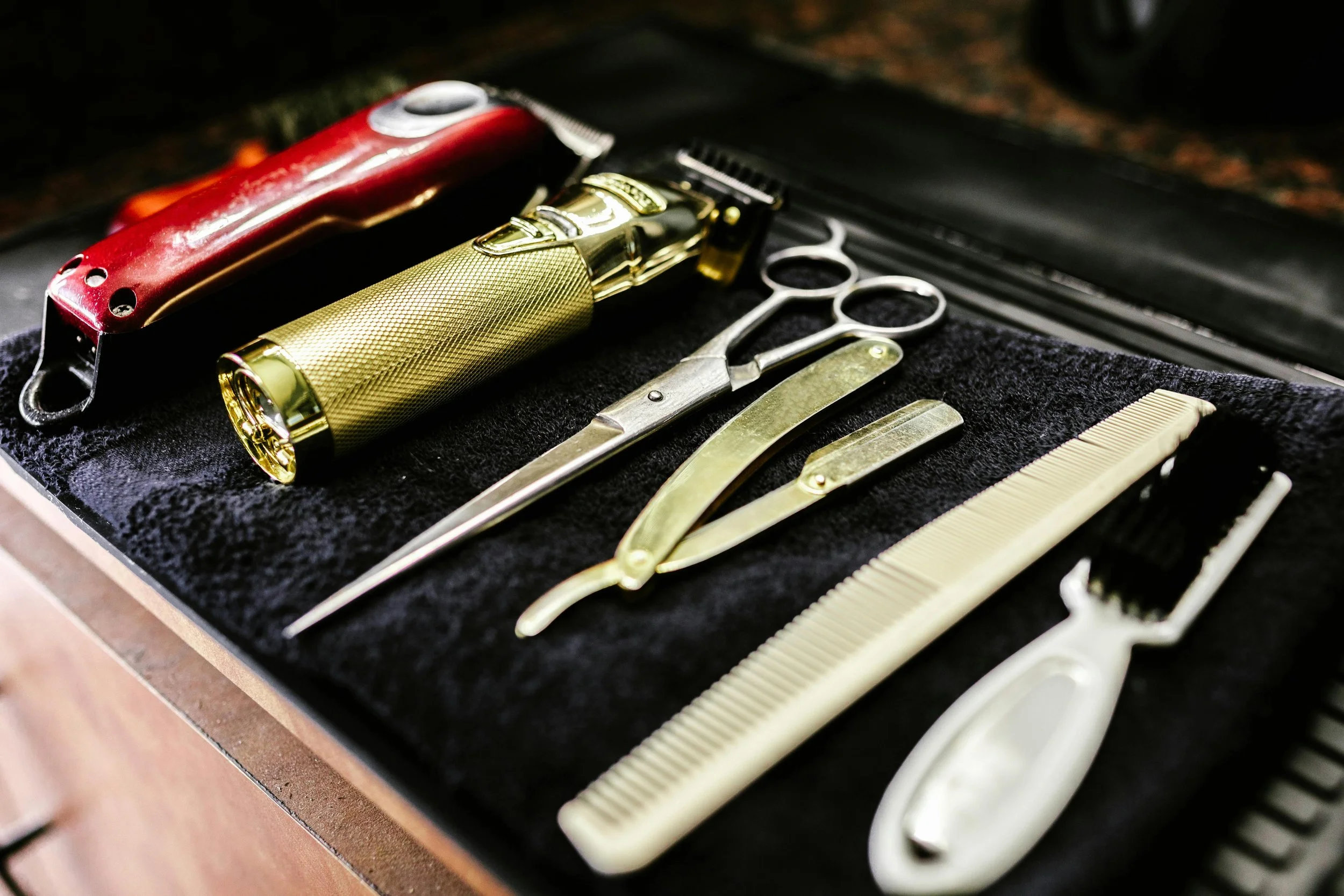 A collection of barber tools including a straight razor, scissors, comb, clippers, and a spray bottle, arranged on a black surface.