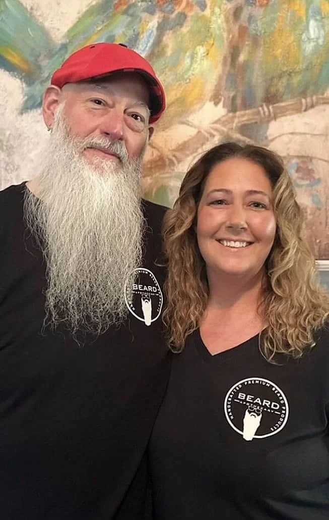 A man with a long white beard wearing a red cap and a woman with curly blonde hair smiling, both wearing black t-shirts with a bearded face logo and the word 'BEARD' in a circular design. They are standing indoors in front of a colorful, abstract mural.