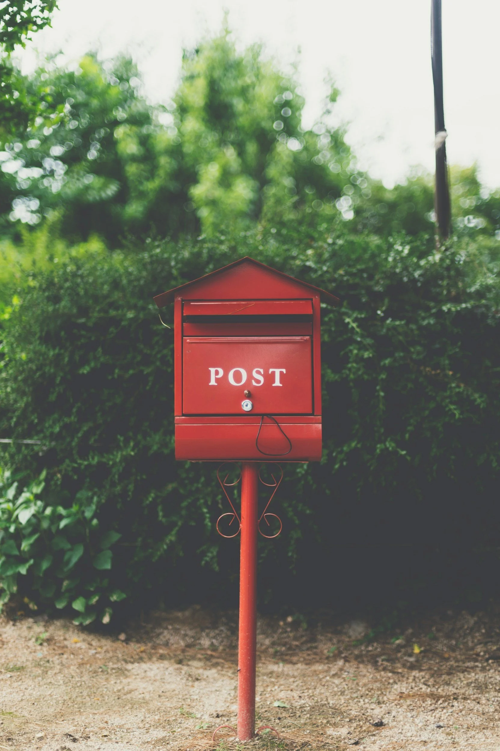 Red mailbox labeled 'POST' mounted on a metal pole in front of green shrubbery.