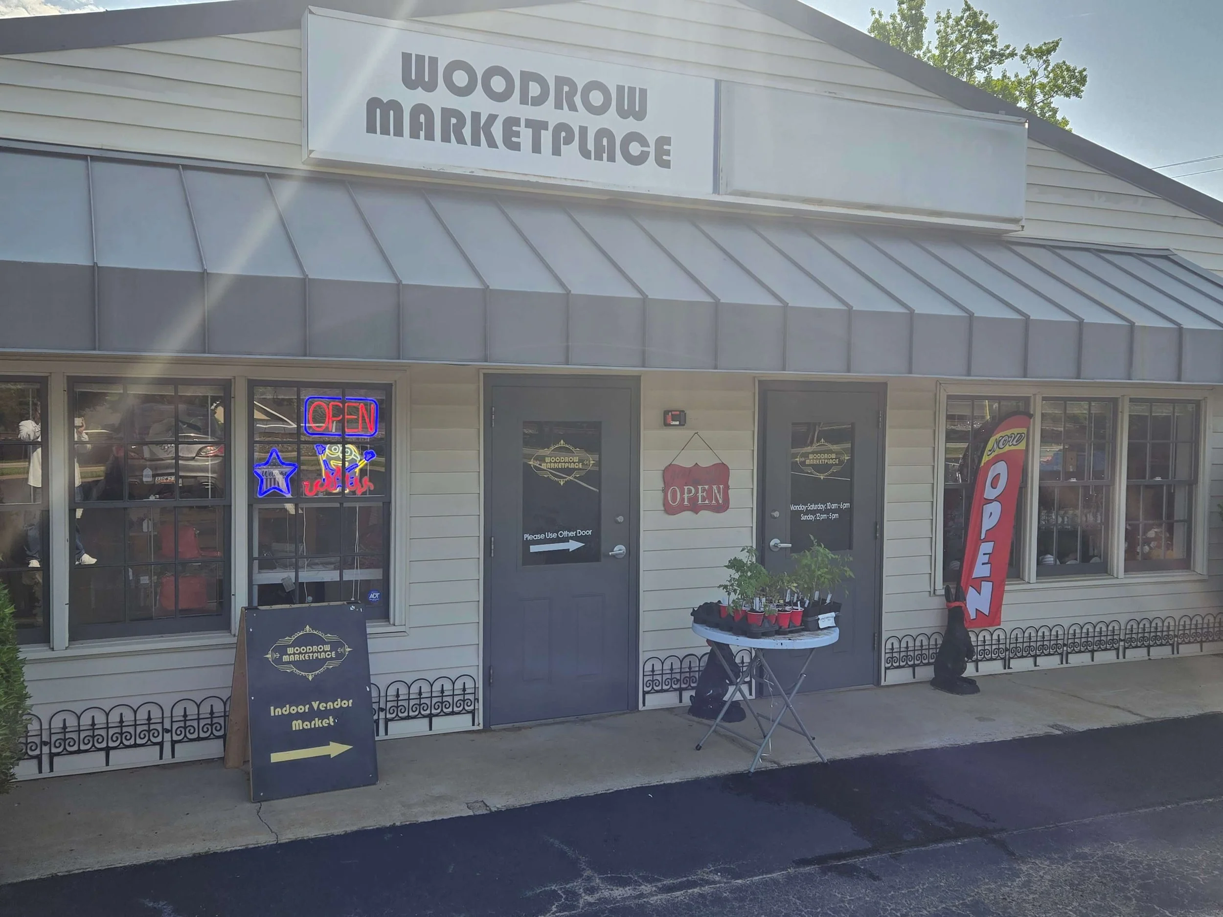Exterior of a storefront with a sign reading 'Woodrow Marketplace.' The building has windows and doors, with neon signs that say 'Open', 'Wild', and a star. There is a blackboard sign indicating 'Indoor Vendor Market' with an arrow, a table with potted plants in front, and a red vertical flag with the word 'Open' on it. The sky is clear and the parking lot is visible.
