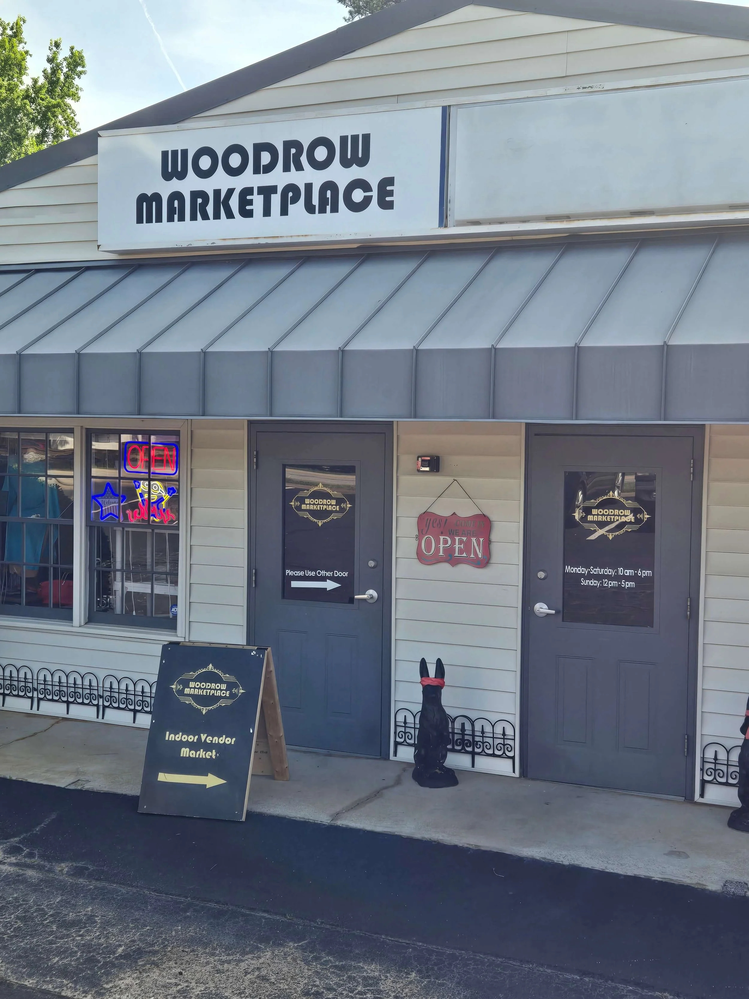 Exterior of Woodrow Marketplace with a large sign, two doors, a small sidewalk sign pointing to an indoor vendor market, and decorative items including a black dog statue with red accents and a neon sign in the window.