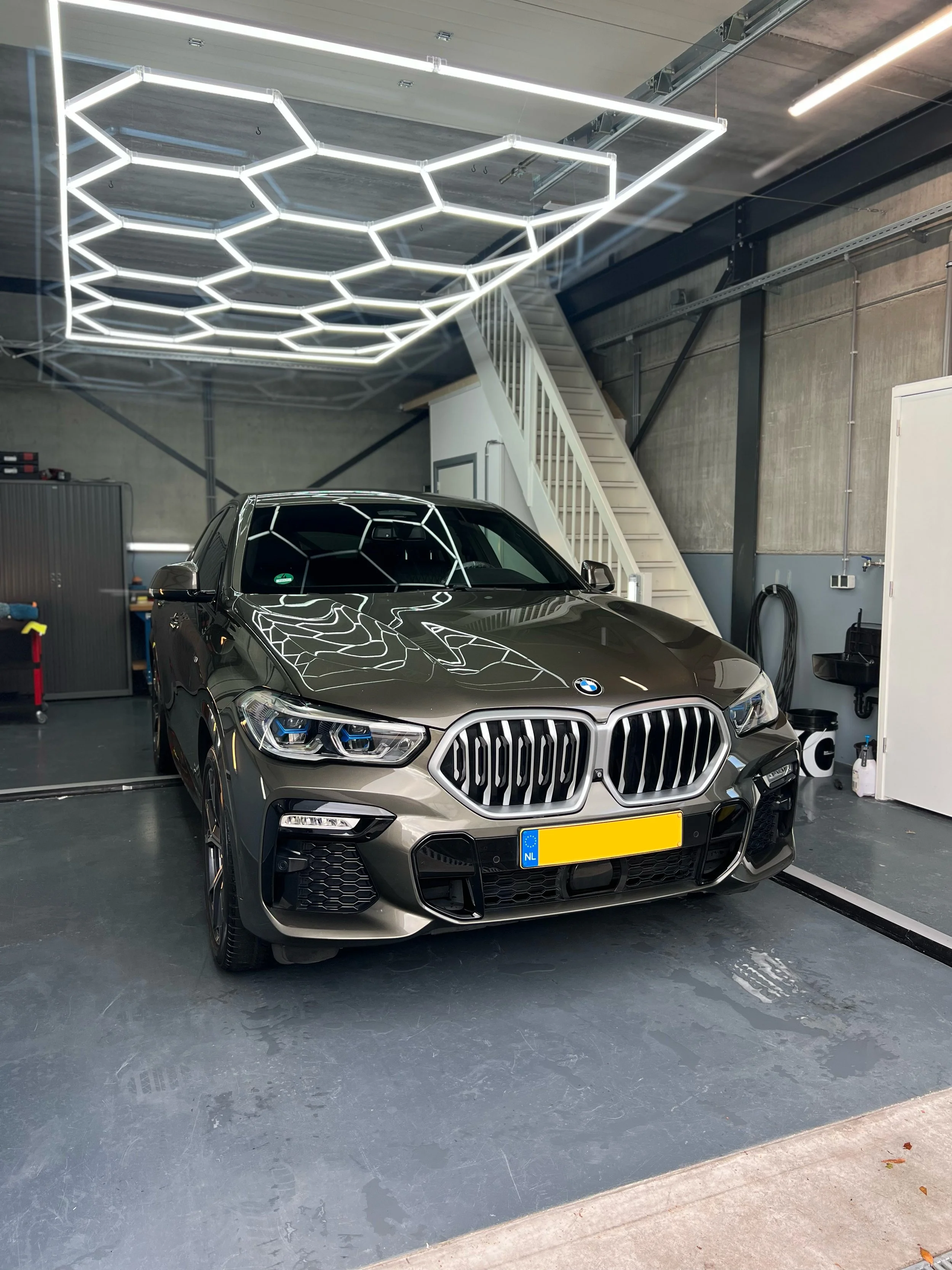 A BMW car parked inside a garage with modern hexagon-shaped neon ceiling lights reflected on the car's hood and windshield.