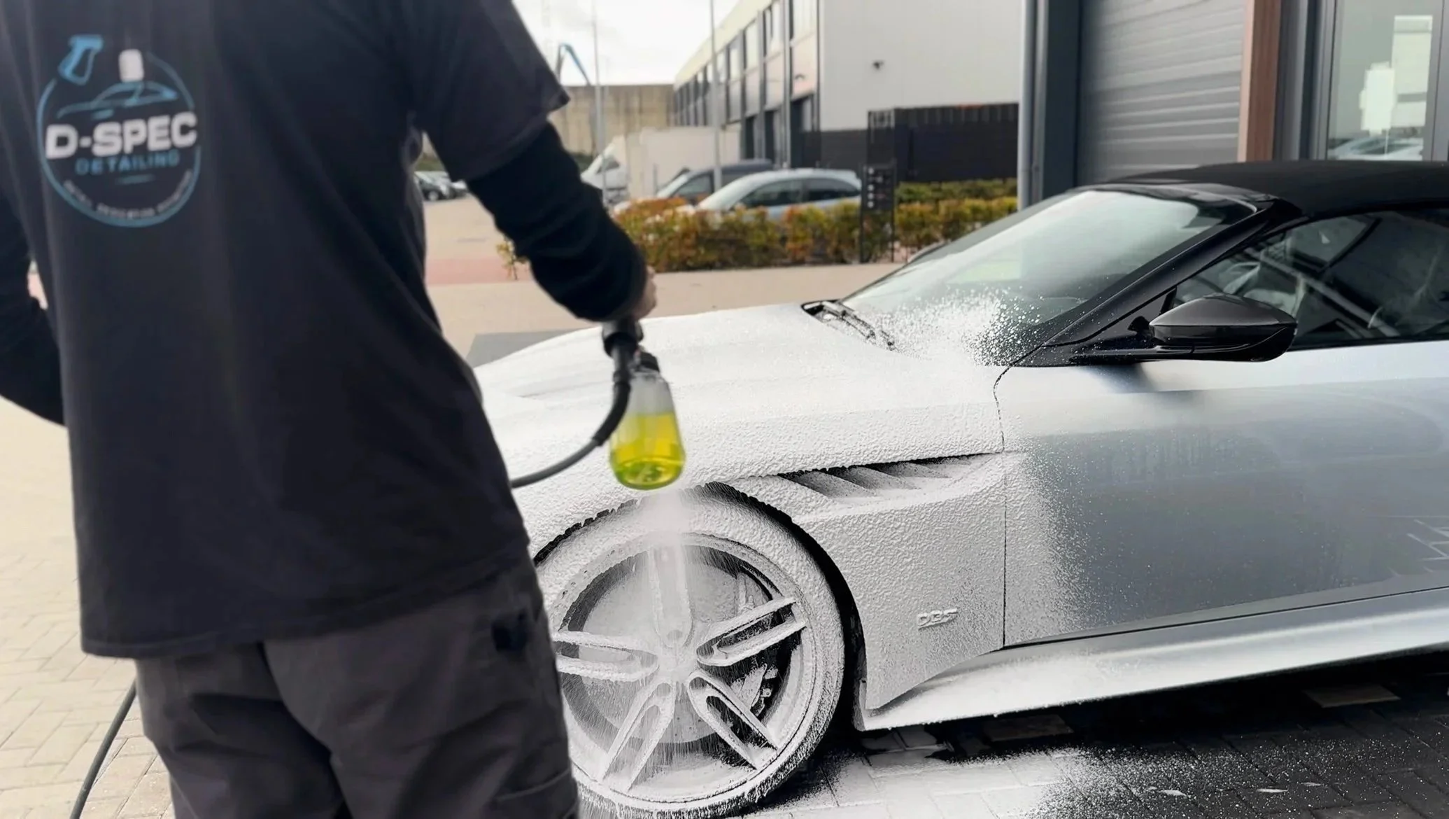 A person pressure washing a silver sports car covered in foam at an auto detailing shop.
