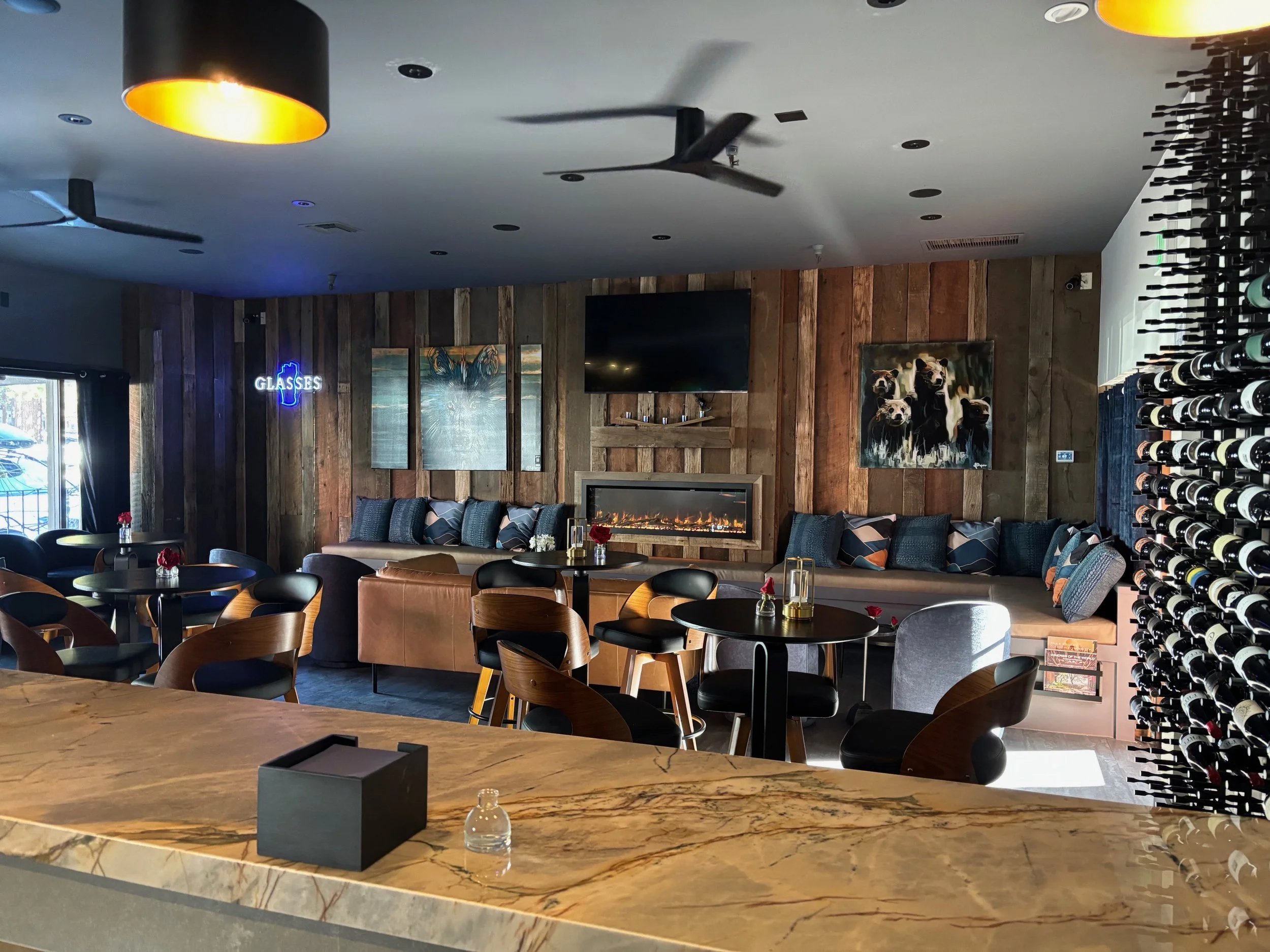 Interior of a modern stylish bar or lounge with a marble countertop, black tables, and chairs, a wooden accent wall with paintings and a mounted TV, a neon sign that says 'GLASSES', and a wine rack on the right side.