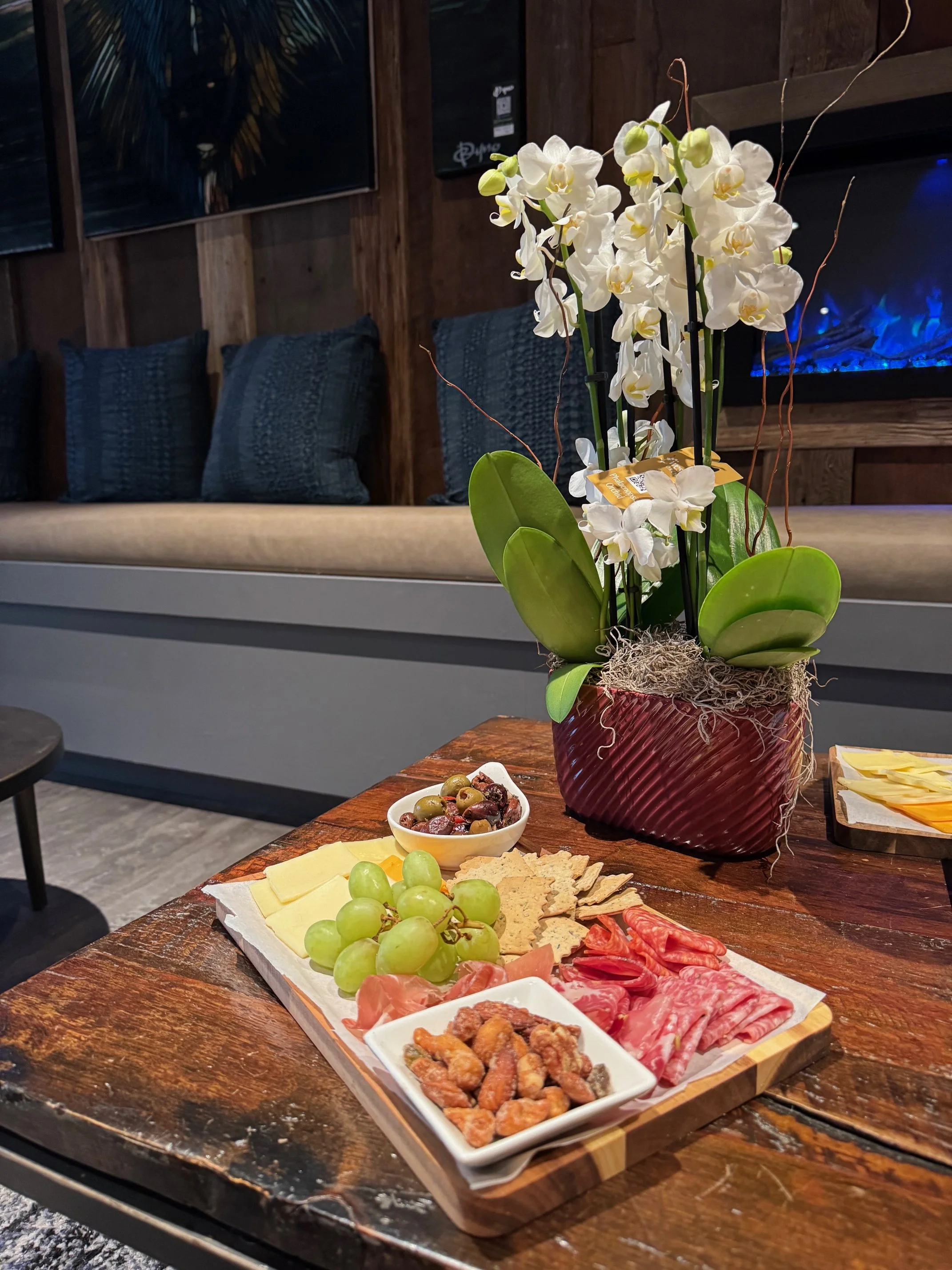 Charcuterie board with grapes, cheese, crackers, cured meats, olives, and a bowl of mixed olives, next to a potted white orchid on a wooden table.