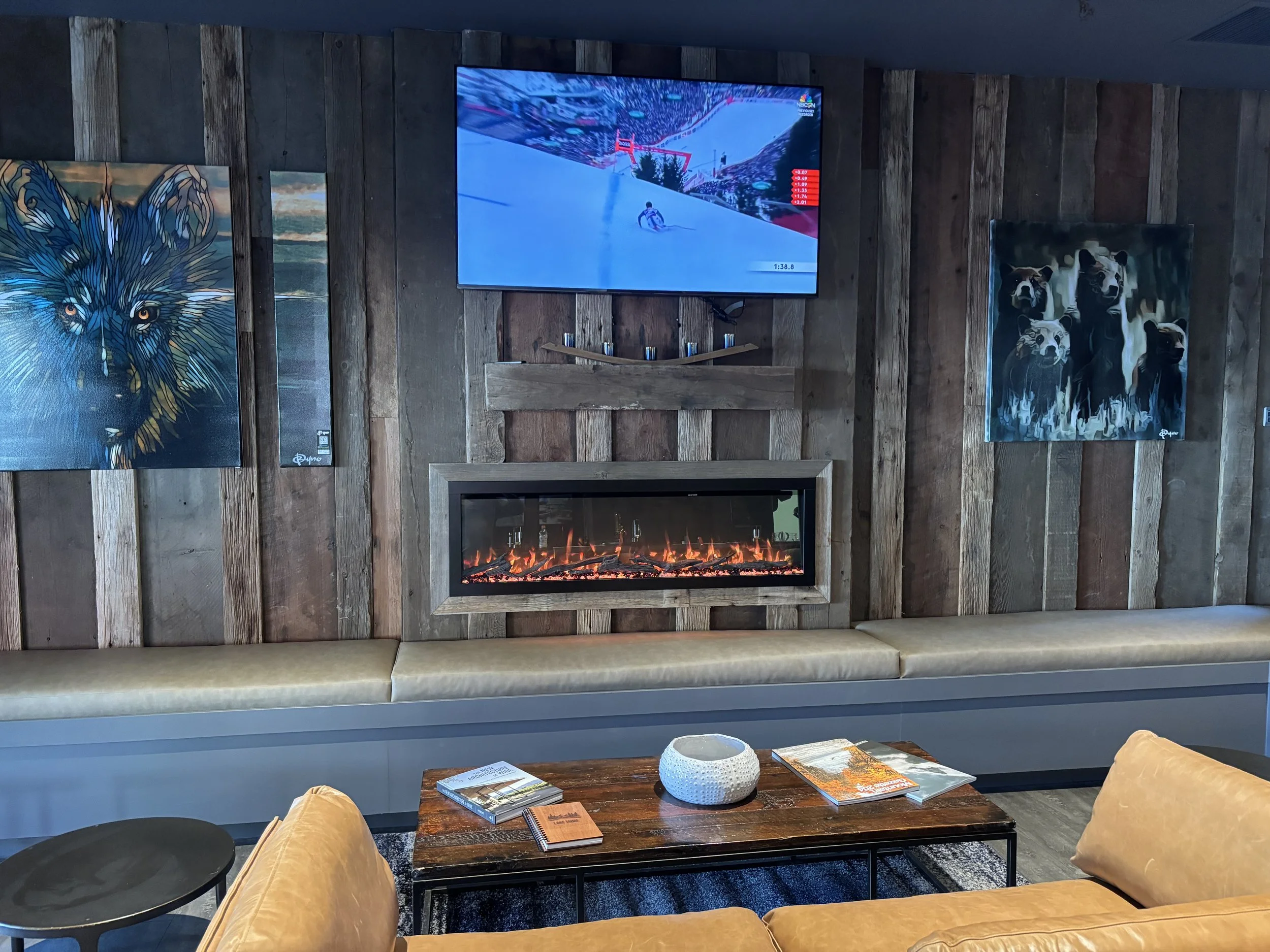 Living room with a mounted TV showing a snowboarding scene, a modern fireplace underneath, and art paintings of a wolf and multiple dogs on the wooden-paneled wall.