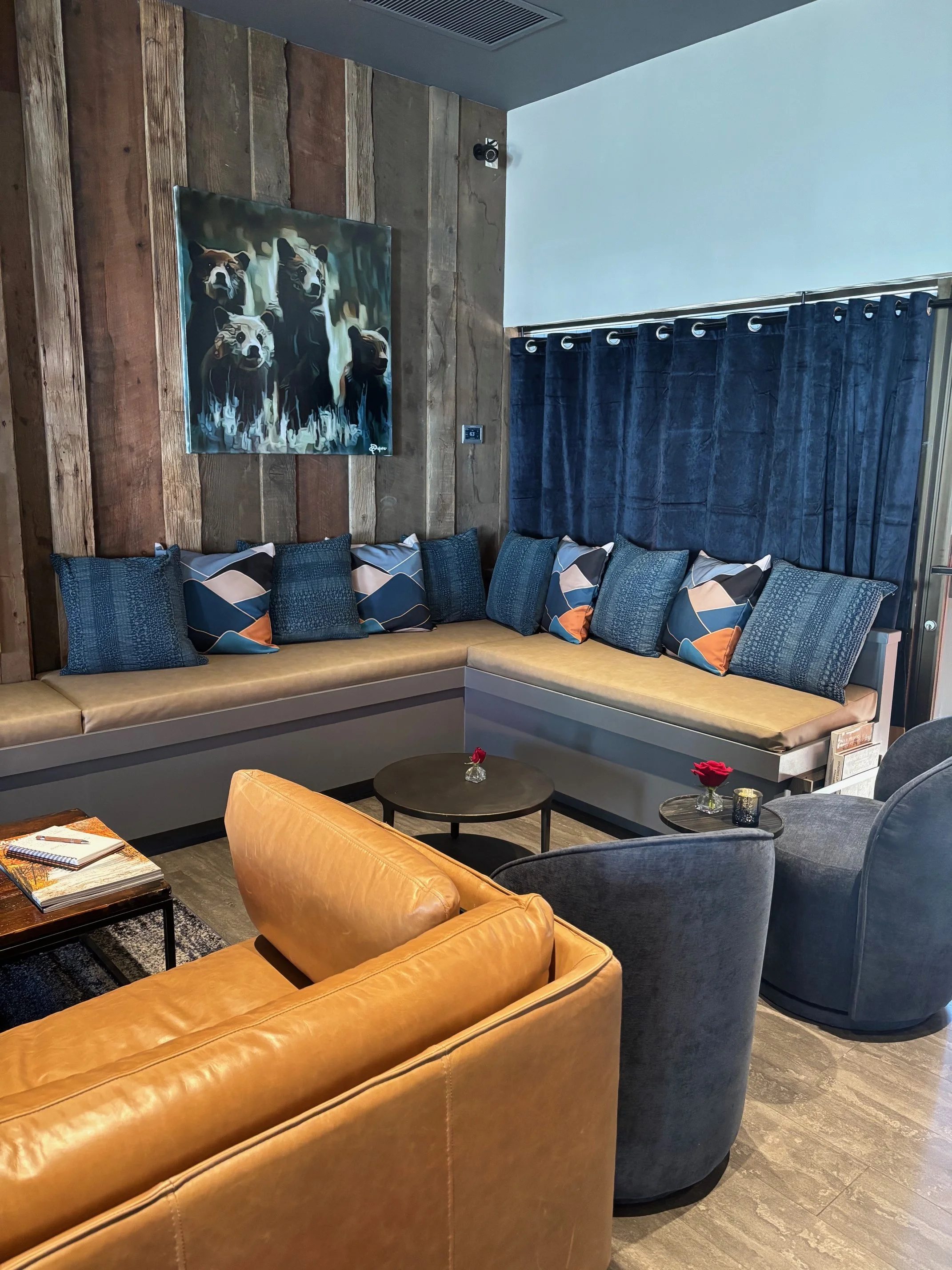 A cozy living room with a mustard yellow leather sofa, two gray velvet armchairs, and a built-in blue cushioned bench against a wood-paneled wall. The wall features a painting of five bears, and there are blue curtains covering a window. Two small ro