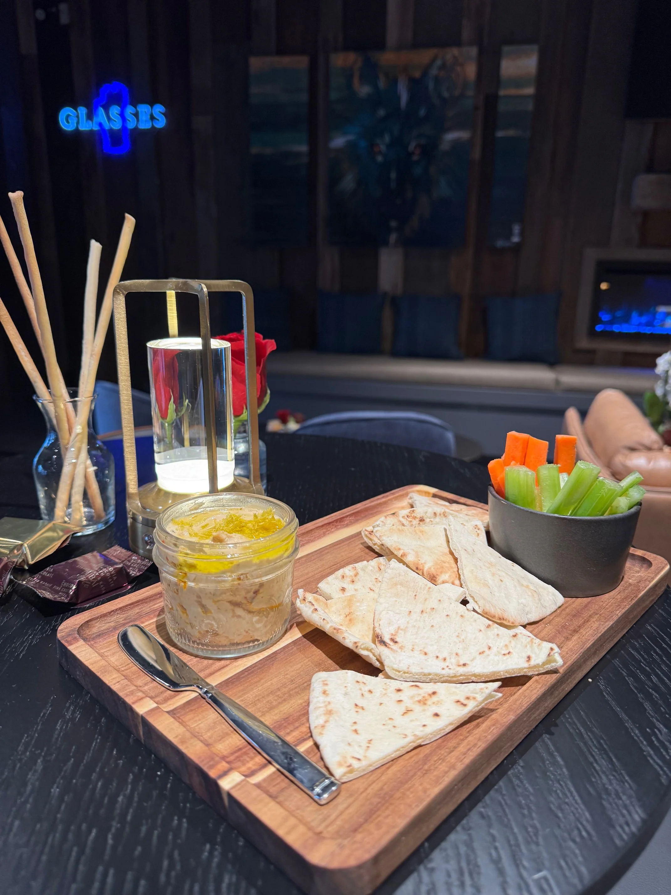 Appetizer plate with flatbread, celery, carrots, hummus, and a candle in a jar on a wooden board in a restaurant setting.