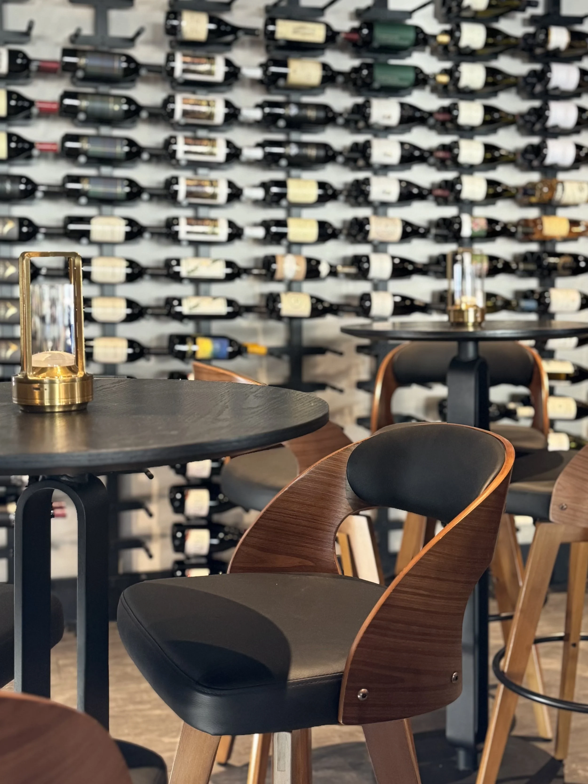 Interior of a wine bar or restaurant with modern wooden chairs with black cushions, round black tables, and a large wall-mounted wine rack filled with bottles in the background.