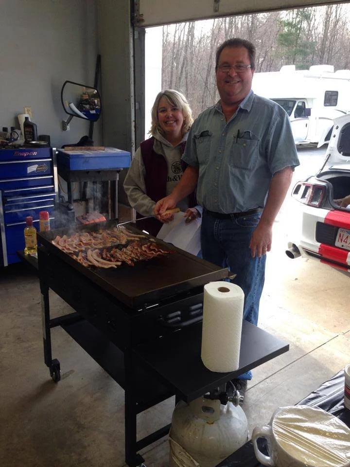 Two smiling people, a woman and a man, standing next to a grill with bacon cooking on it. They are inside a garage with various tools and a mirror on the wall, and a large window showing an RV and trees outside.
