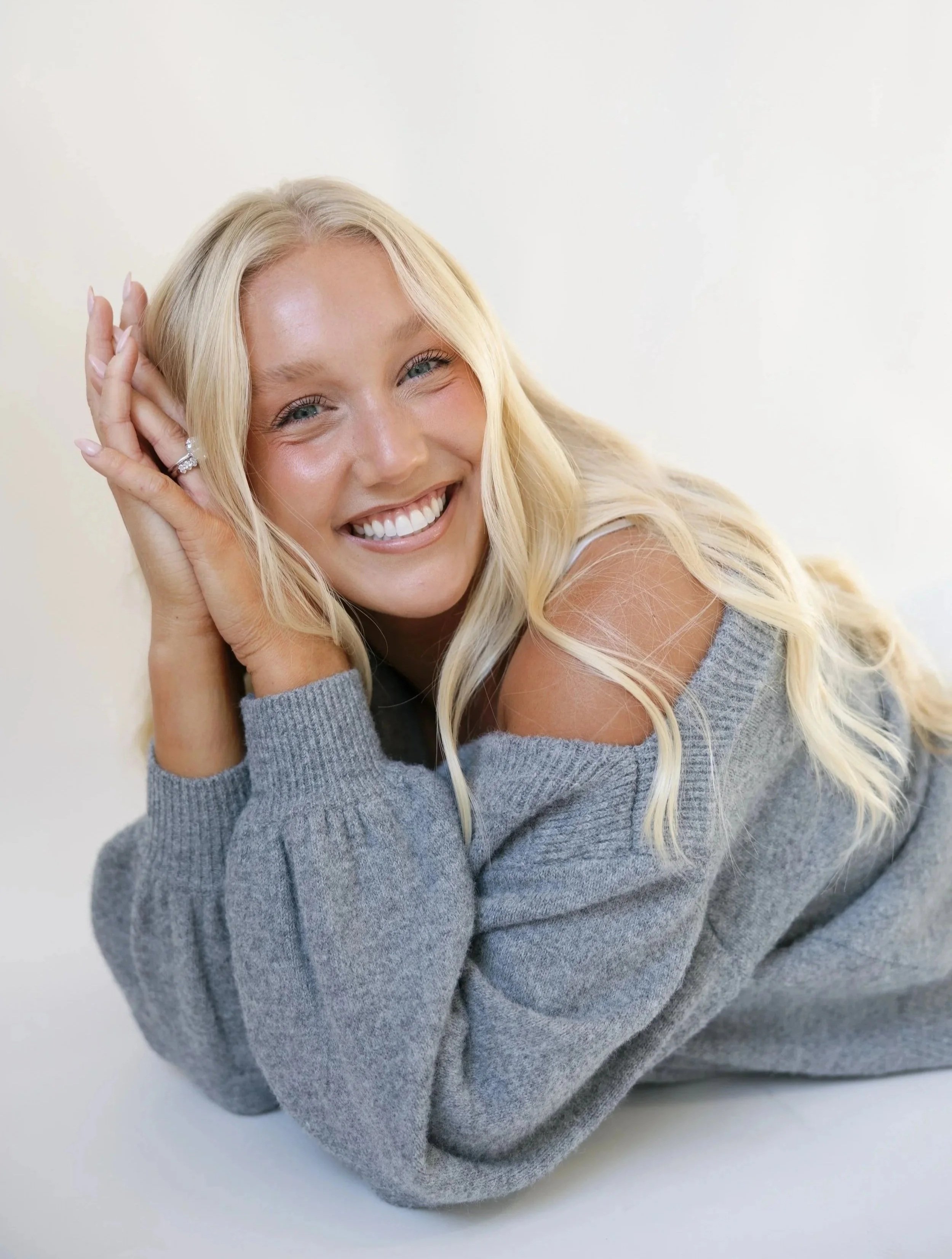 A smiling blonde woman in a gray sweater, lying down with her hands near her face, looking at the camera.