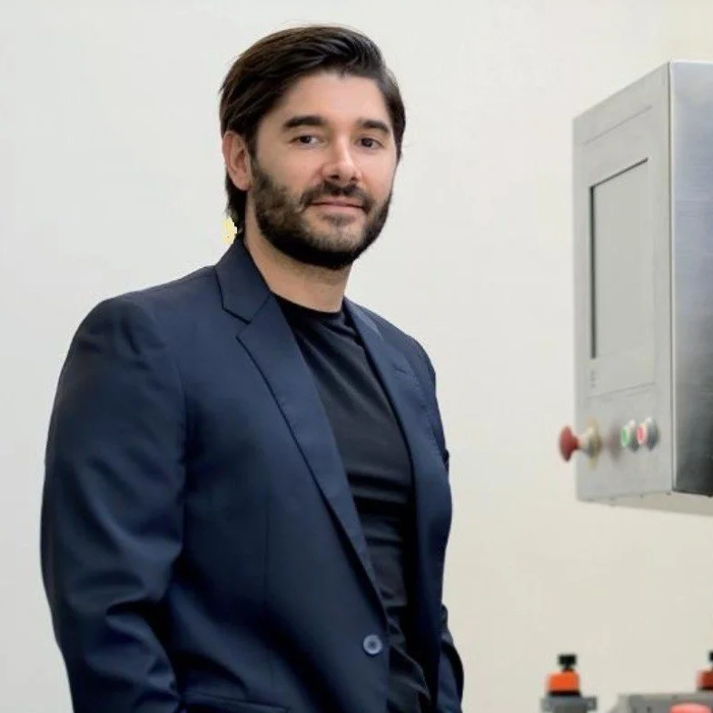 A man with dark hair and a beard, wearing a navy blazer and black shirt, standing next to a stainless steel machine with buttons and a small display, against a plain light-colored wall.