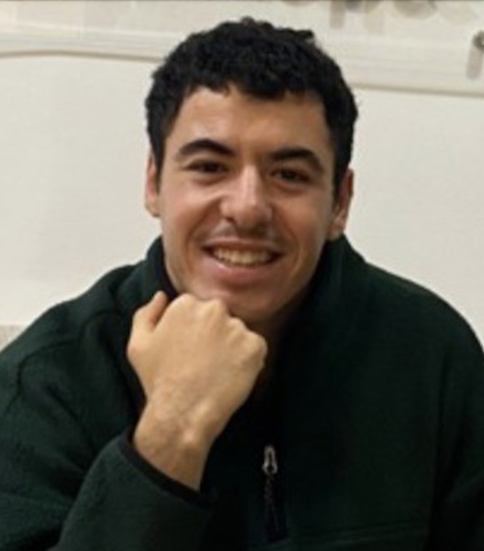 A young man with dark curly hair smiling, resting his chin on his hand, wearing a dark jacket, in an indoor setting.