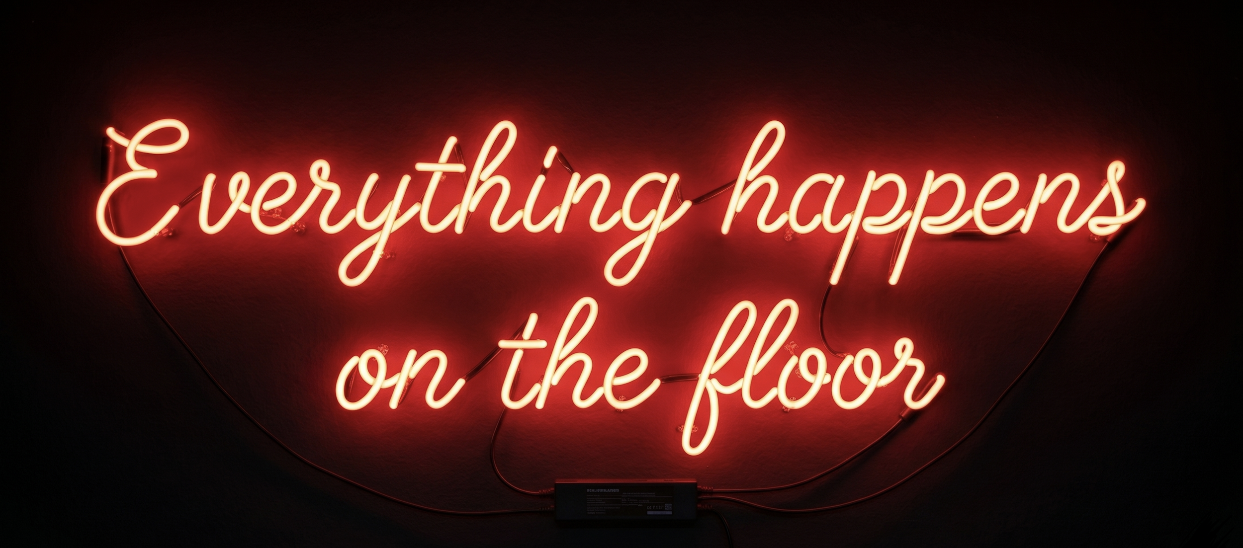 Neon sign reading 'Everything happens on the floor' in red, mounted on a dark wall.