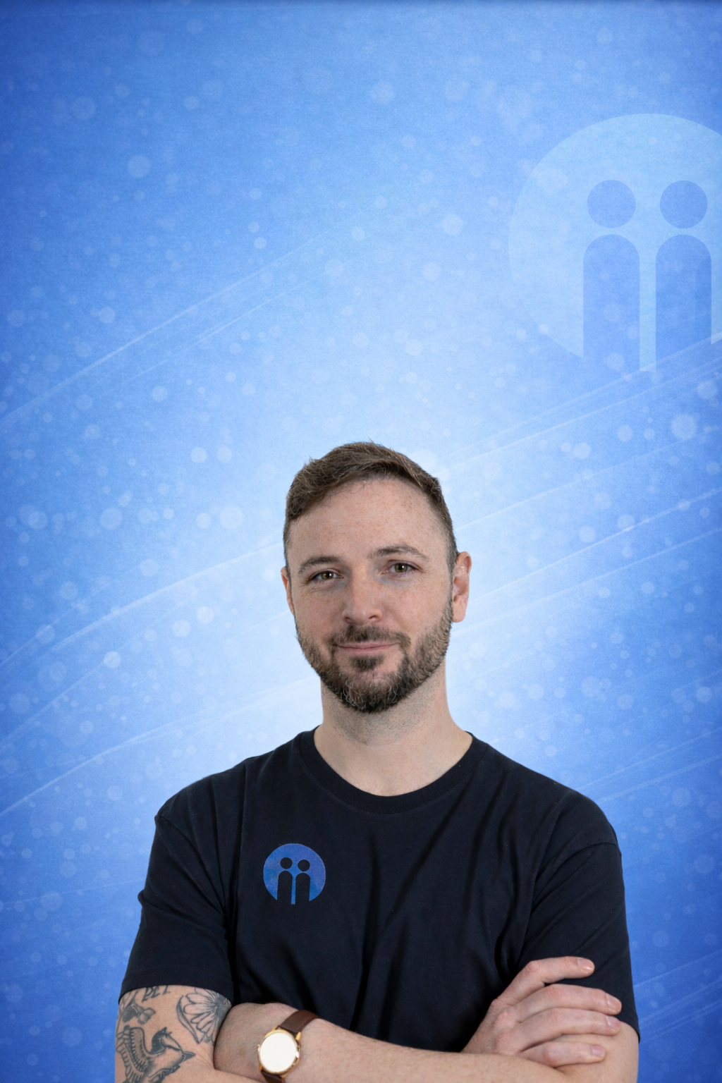A young man with short brown hair and a beard, wearing a black t-shirt with a blue logo, stands in front of a blue background with a pattern of light circles and lines. He has his arms crossed, and is wearing a watch and has tattooed arms.