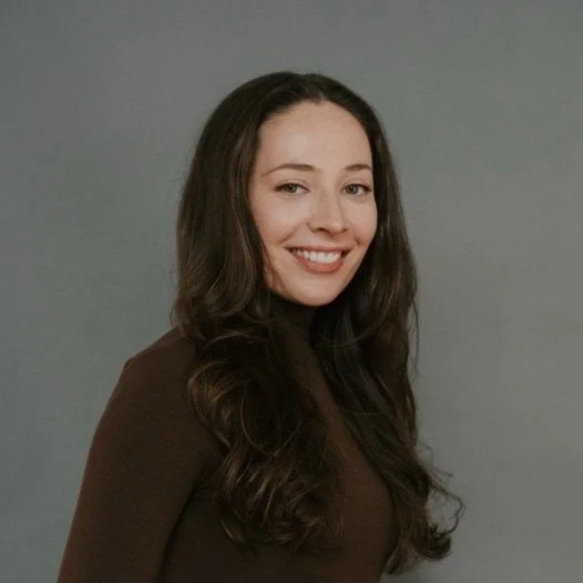 A young woman with long wavy brown hair smiling, wearing a dark long-sleeve top, against a gray background.