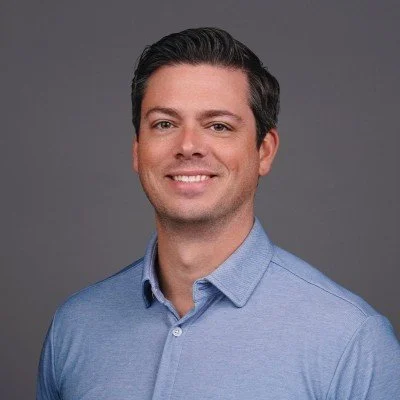 A man with dark hair wearing a light blue button-up shirt smiling against a plain gray background.