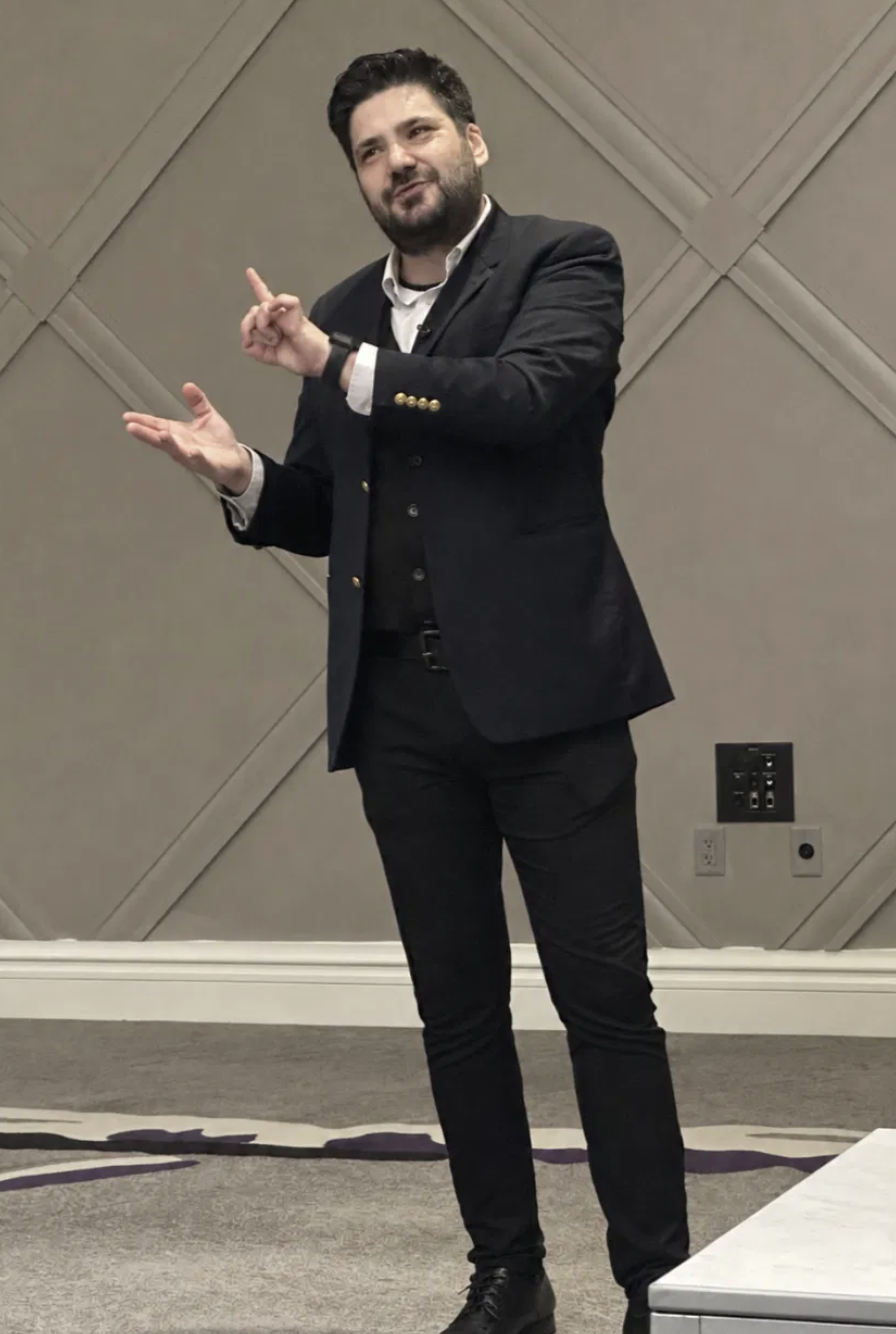 A man with dark hair and a beard, wearing a dark suit and white shirt, gesturing with his hands while speaking at an indoor event.