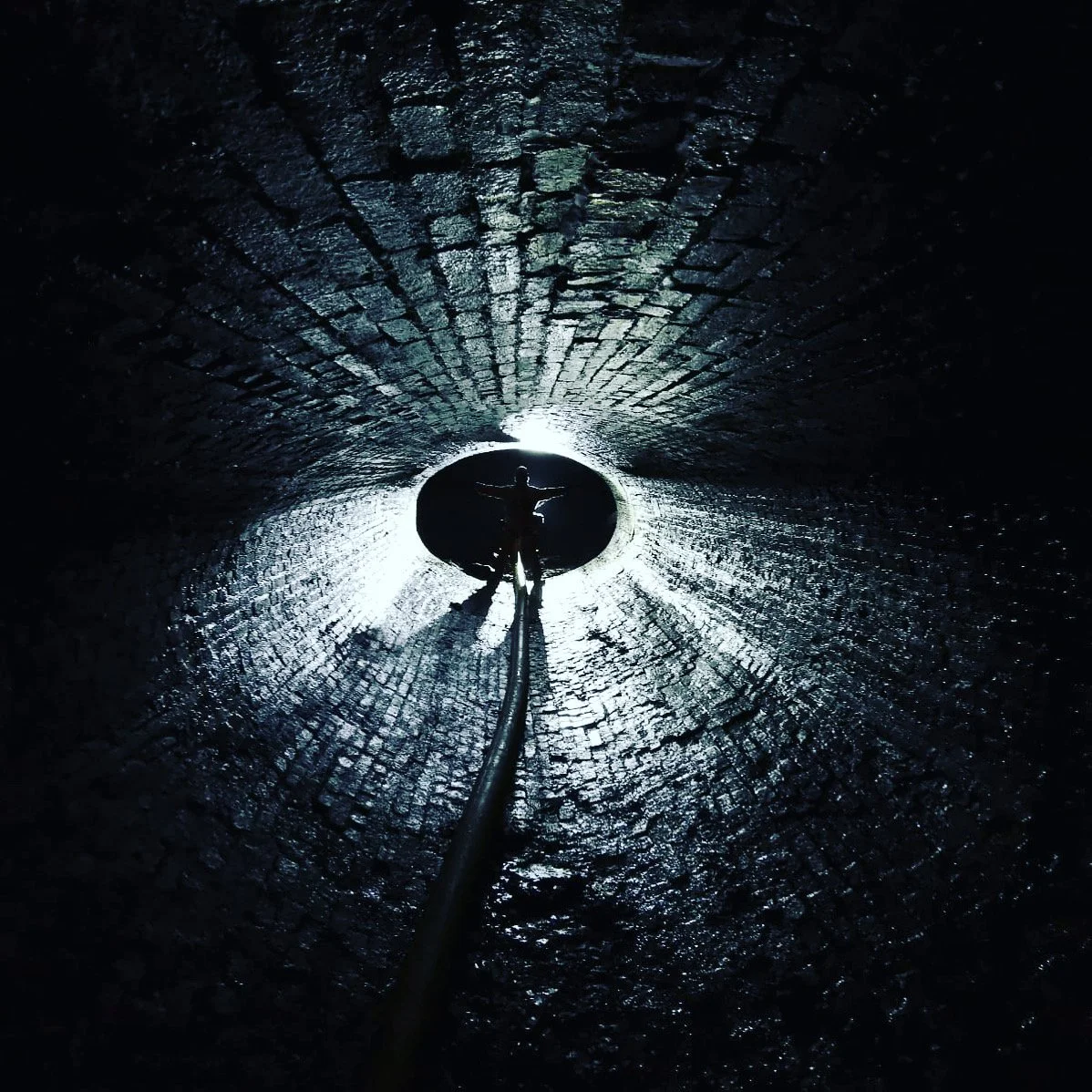 A silhouetted person standing inside a dark tunnel opening to the outside, with arms outstretched and a rope extending towards the camera.