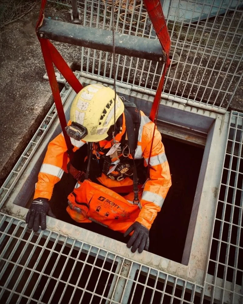 A worker in safety gear, including a helmet and orange reflective jacket, entering a manhole on a construction site.