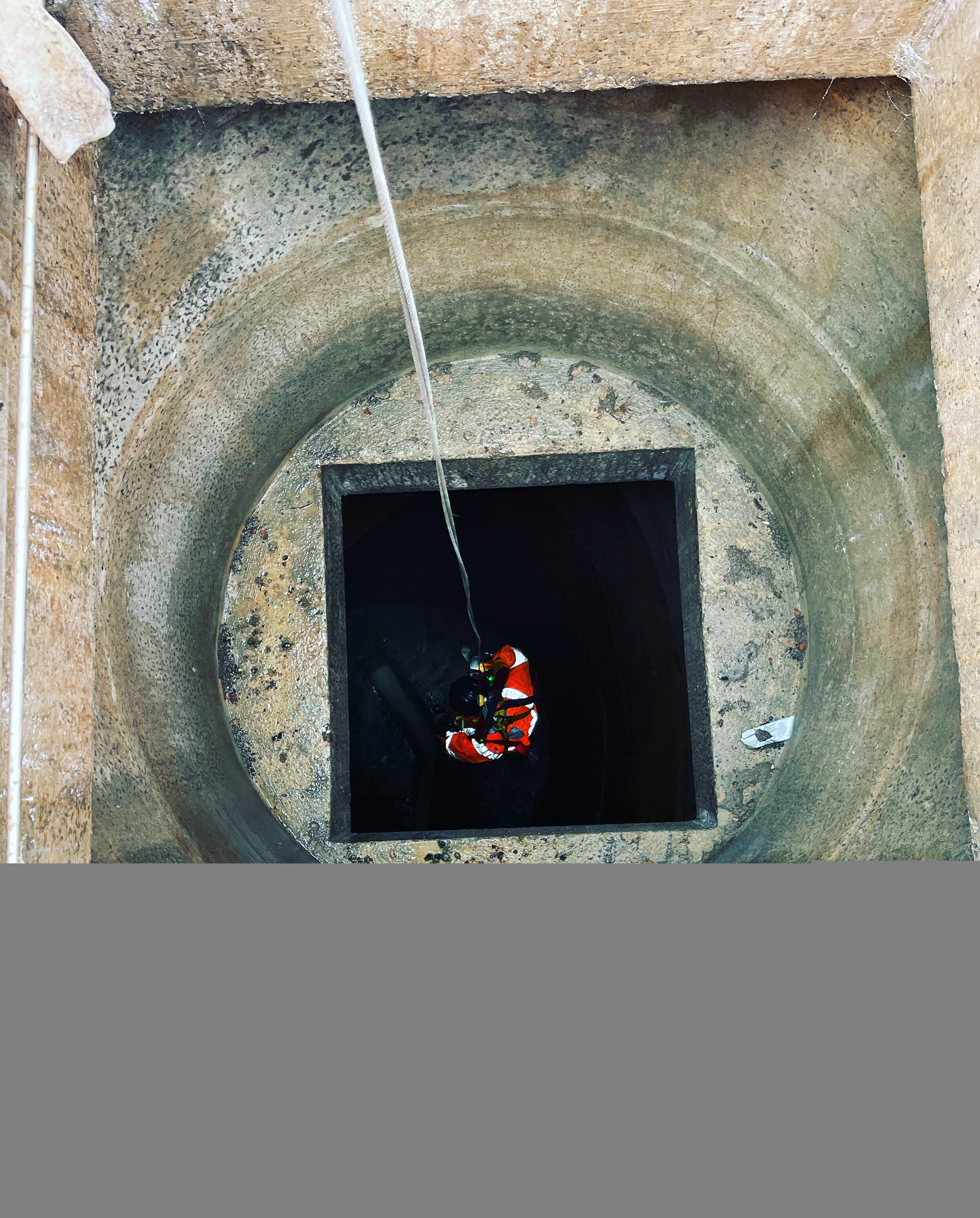 Worker in safety gear using a rope to descend into a large, square, underground concrete structure, viewed from above.