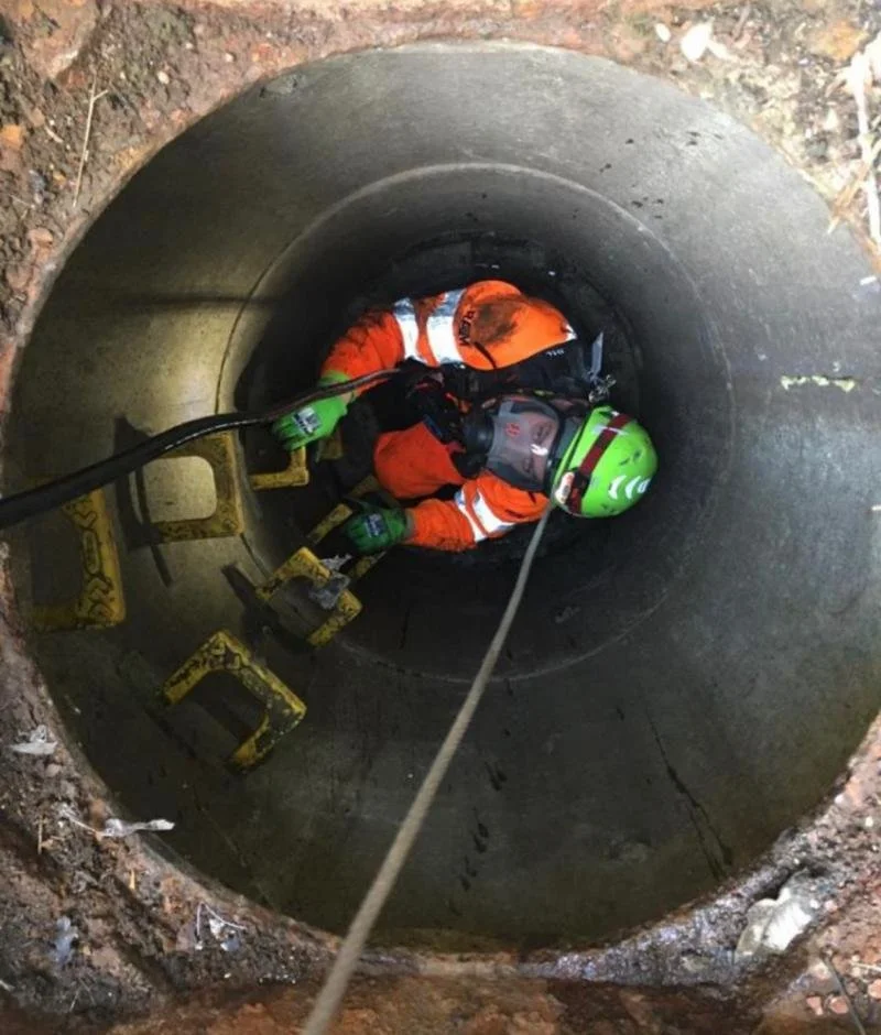 A construction worker in orange safety gear and a green helmet is inside a vertical concrete pipe, using a safety harness and rope for support during a rescue or inspection.