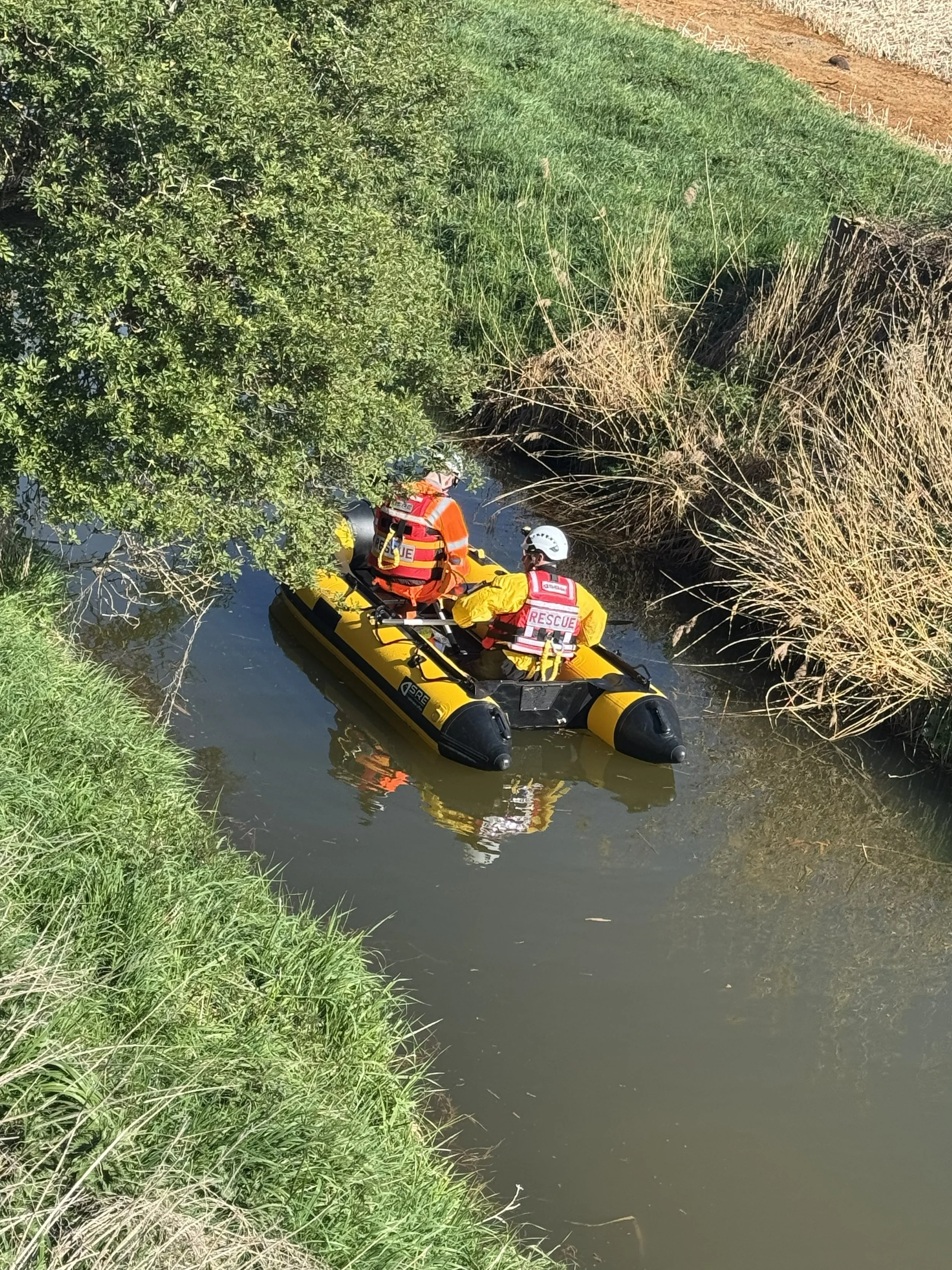 Two rescue workers in an inflatable boat on a narrow waterway surrounded by tall green grass and bushes on the riverbank.