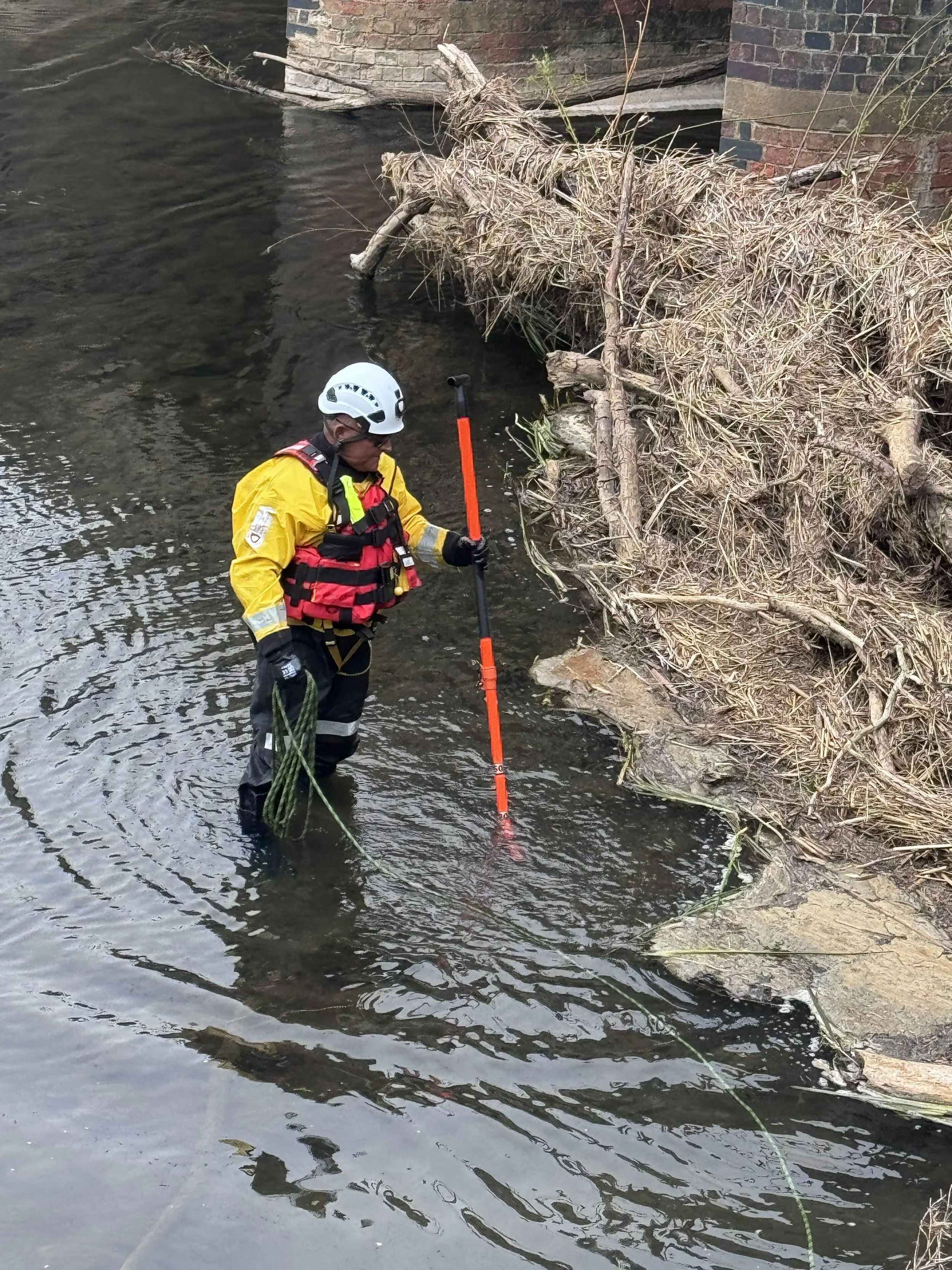 A rescue worker wearing a yellow jacket, a white helmet, and a black face mask stands in a river near fallen debris and dry plant matter. The worker holds a long measuring stick with orange markings, inspecting the water near a brick wall and collaps
