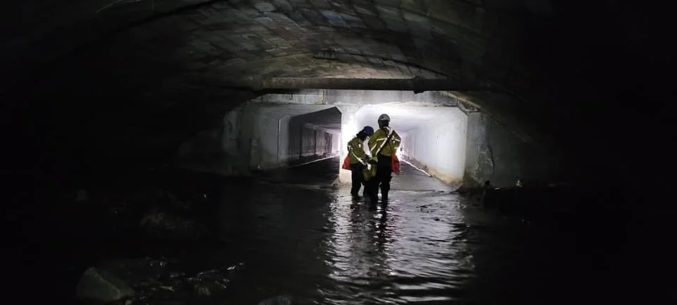 Three workers in safety gear standing in a flooded underpass or tunnel with water reaching their knees, with light shining from the other side.