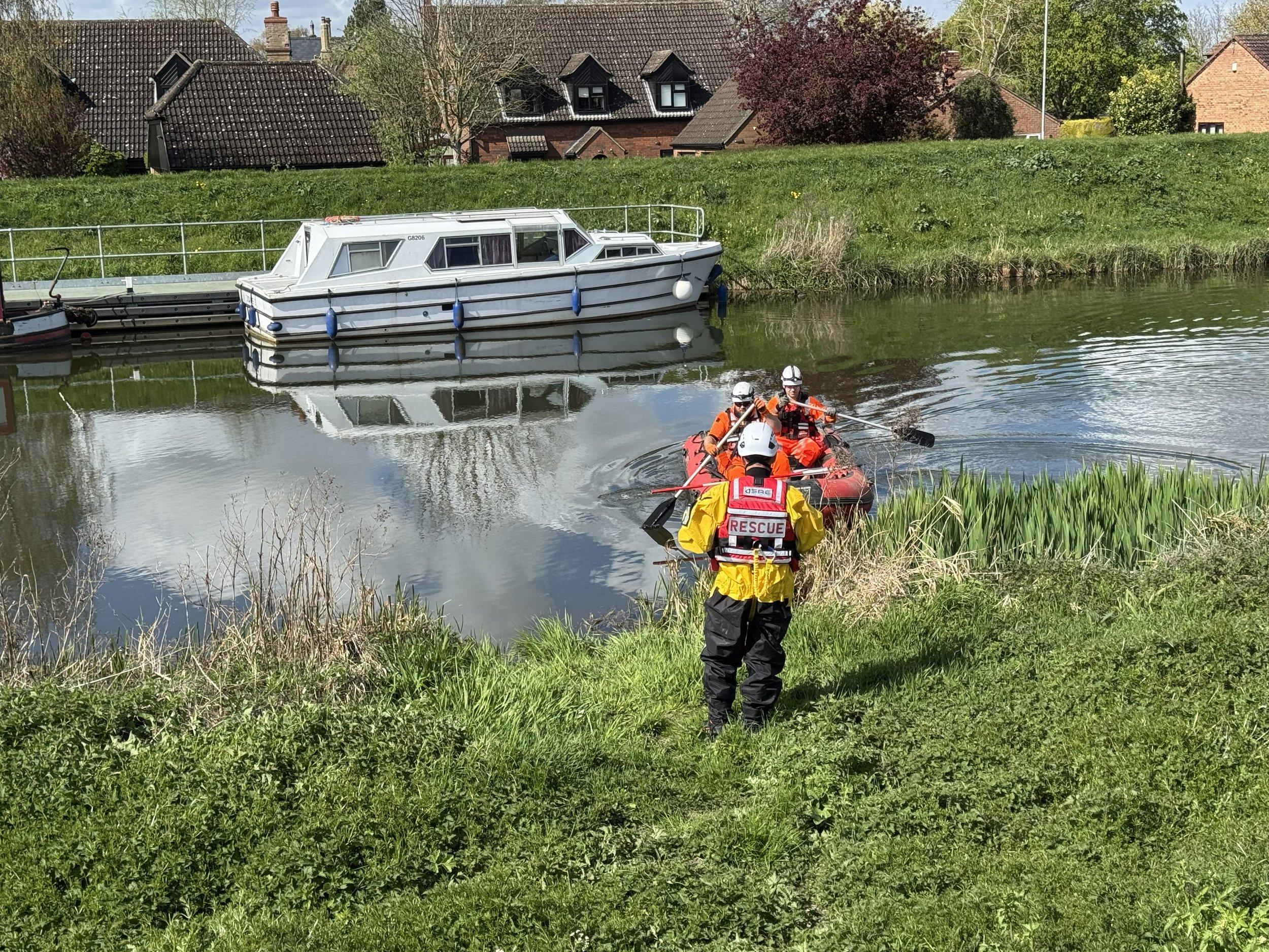 Rescue workers in yellow and orange gear aboard a small red boat on a river, with a house and trees in the background.