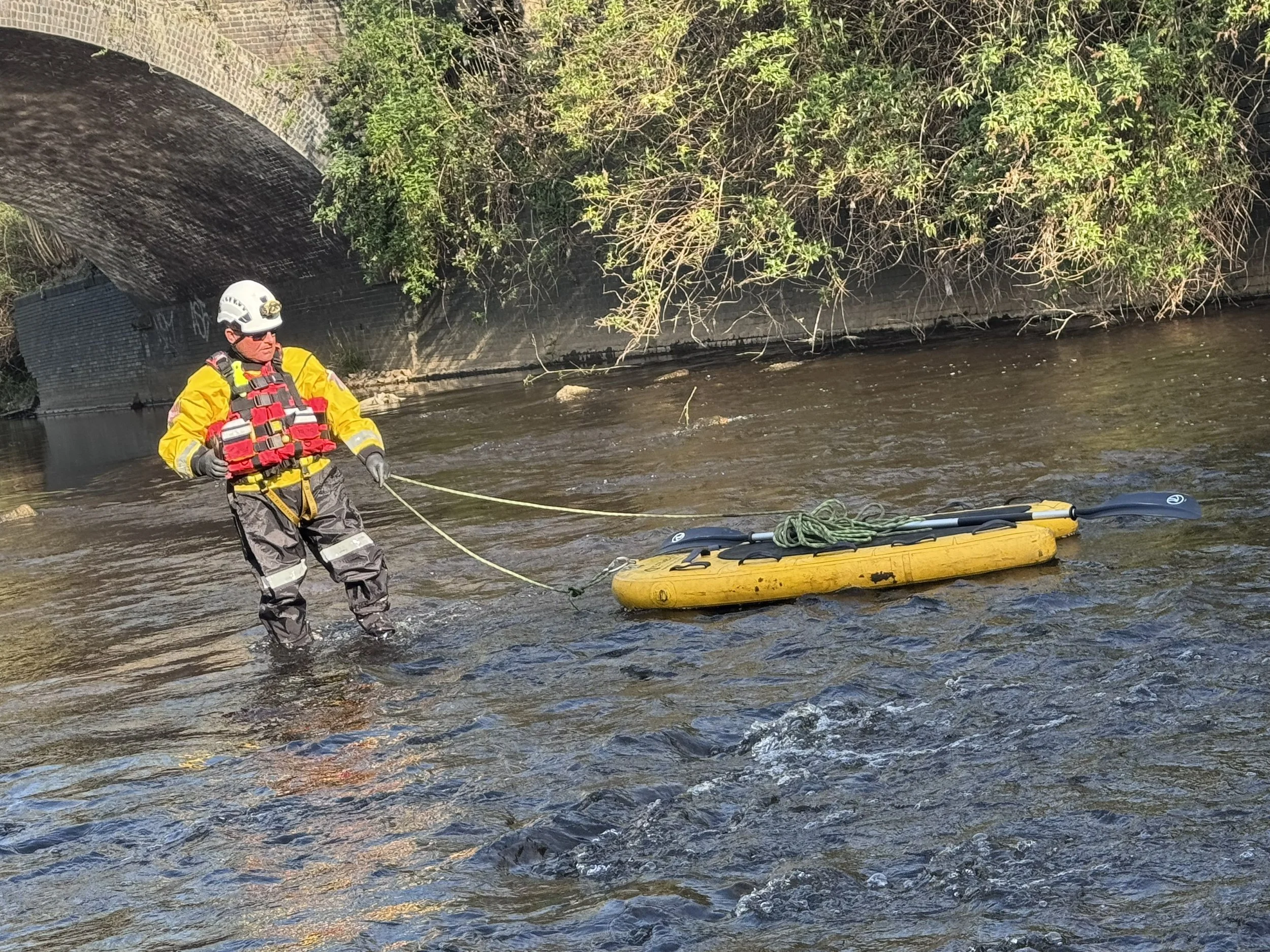 A rescue worker in yellow and gray gear and a white helmet is standing in a river, holding a rope attached to a yellow inflatable boat.