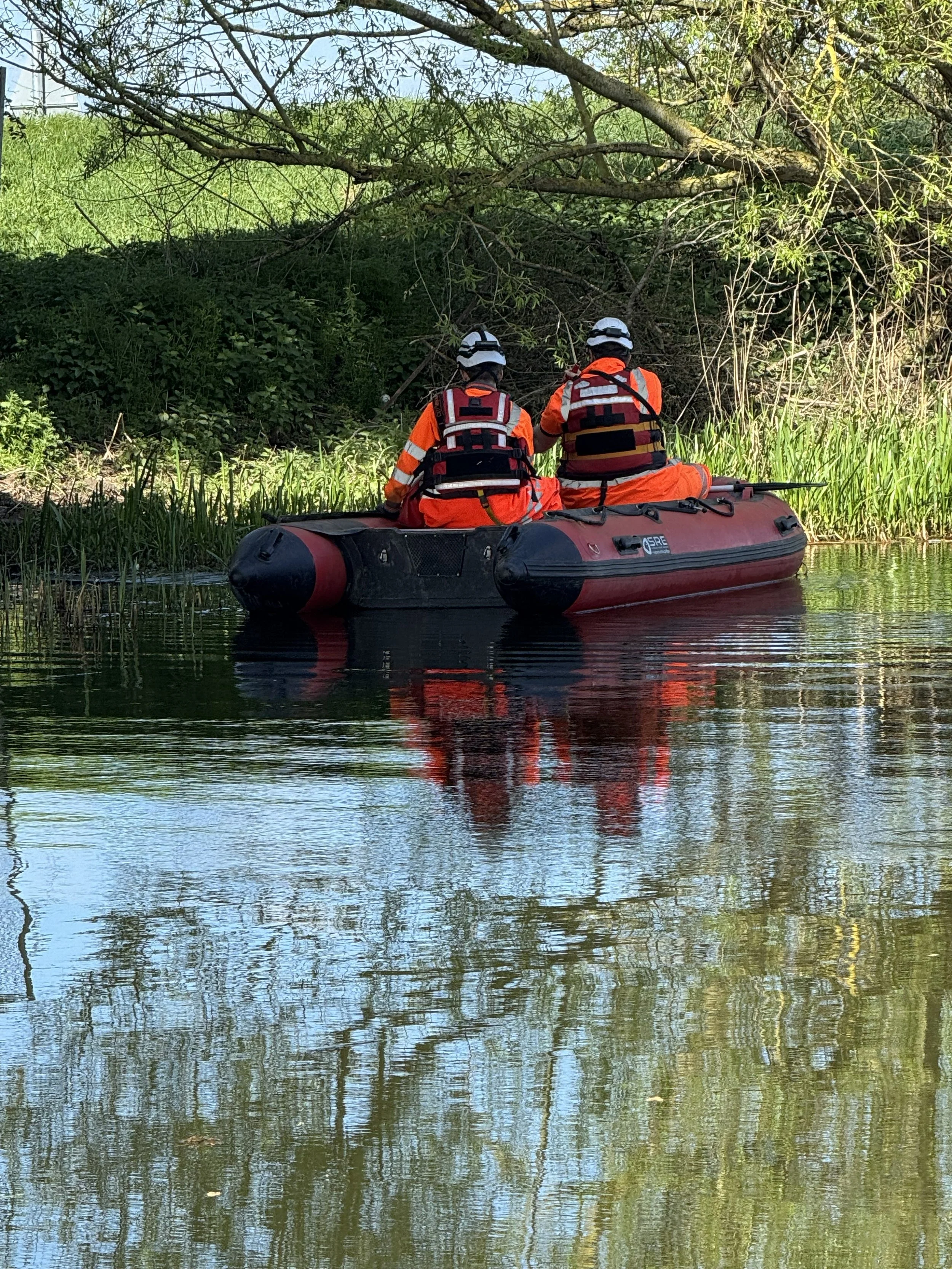 Two rescue workers in orange uniforms and helmets in an inflatable boat on a calm river, near a green, overhanging tree.