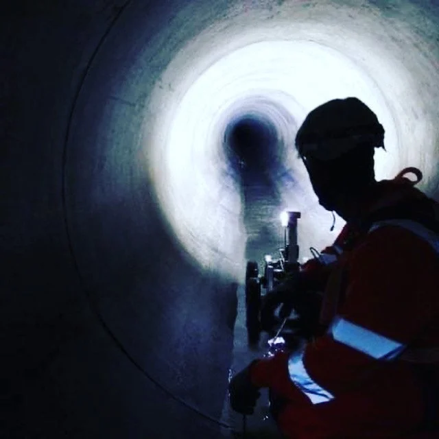 A worker in safety gear and helmet working inside a large, dark pipeline with a bright opening at the end.
