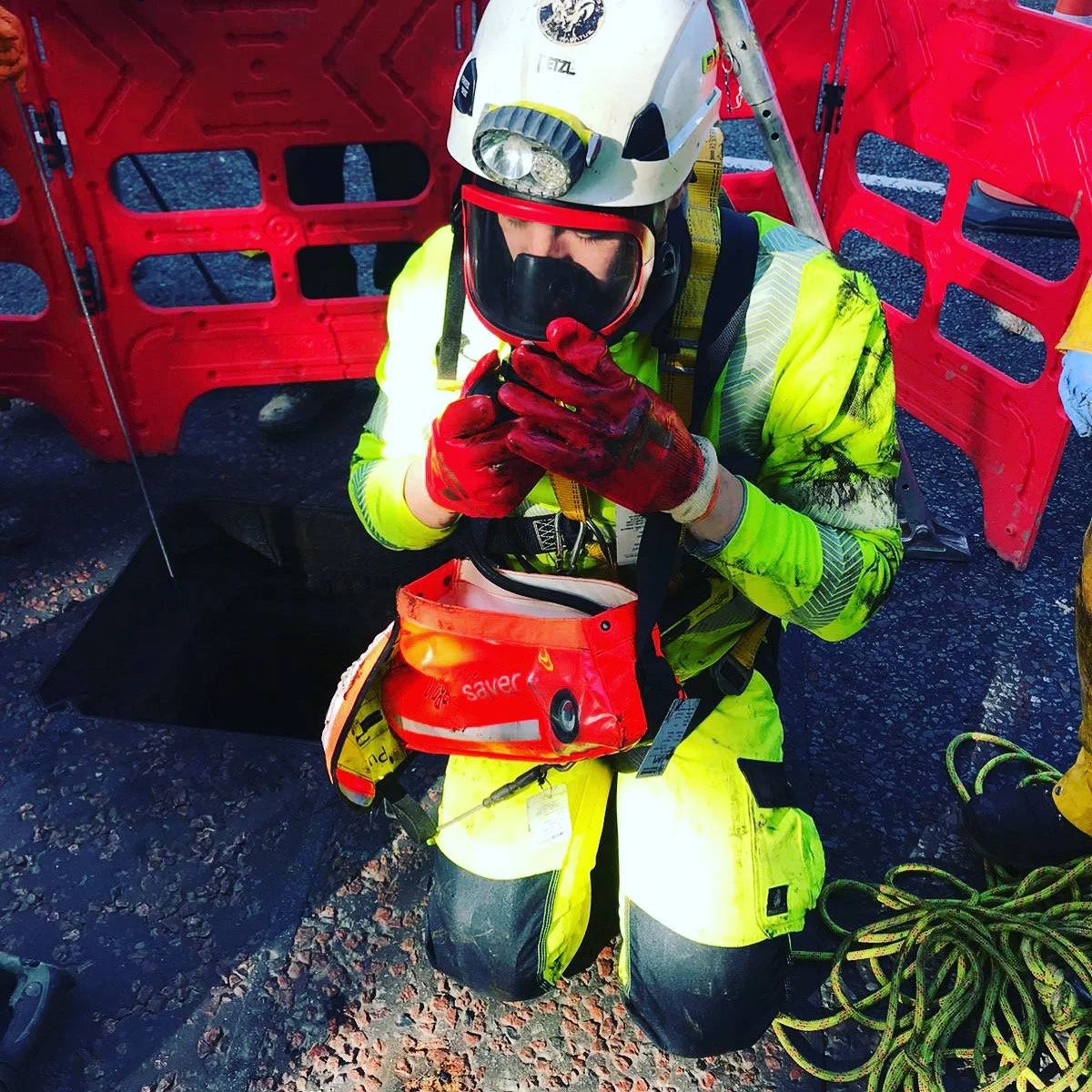 A rescue worker in yellow gear and a helmet with a headlamp kneels on the ground, holding a signaling device with both hands, near a red safety barrier, with climbing ropes nearby, in an outdoor setting.