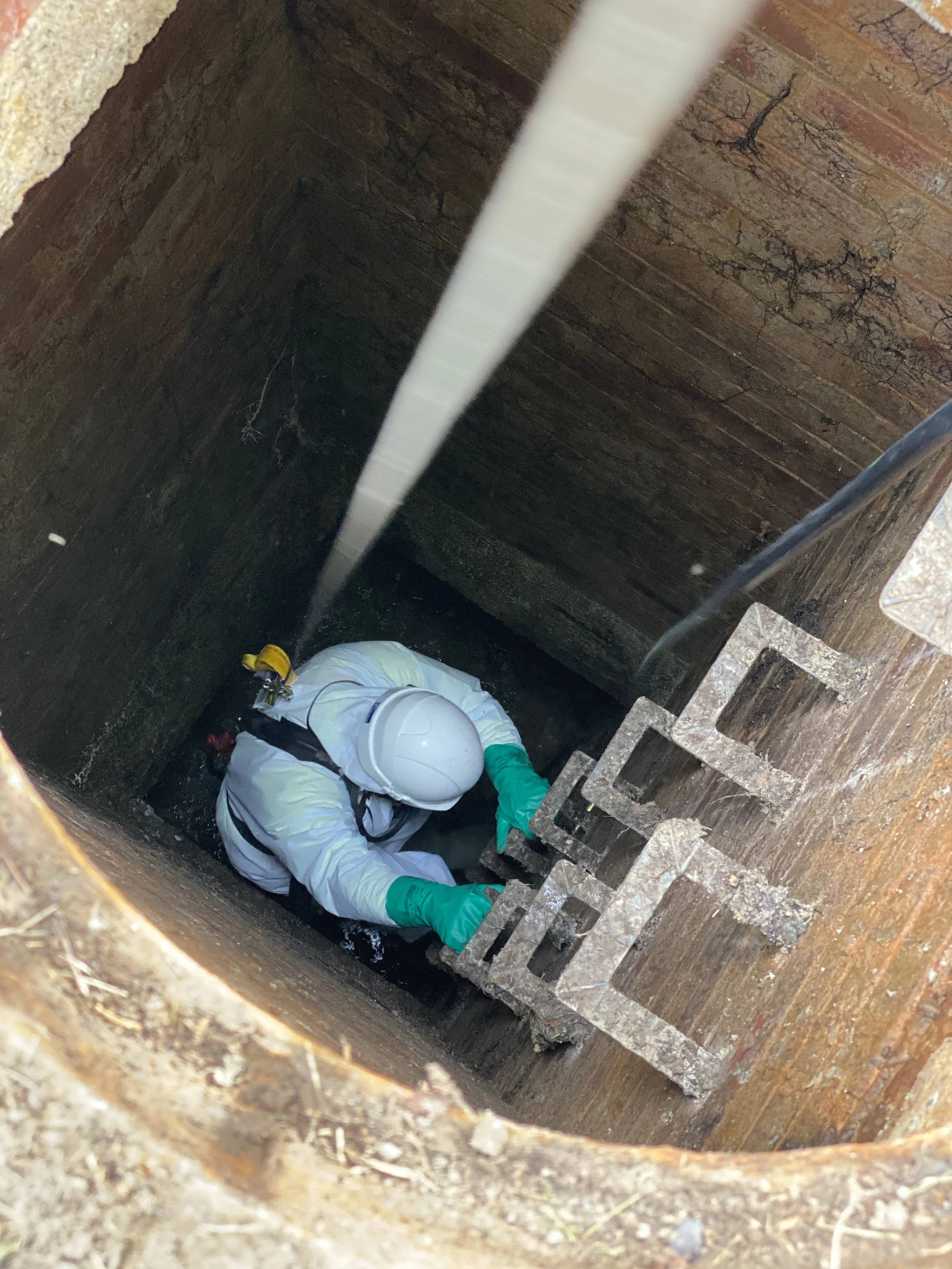 Worker wearing a helmet, protective gloves, and a white suit, descending into a deep, vertical well or shaft with brick walls, using a metal ladder.