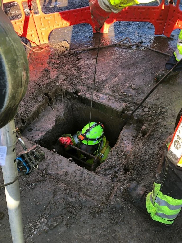 Worker in a safety helmet and vest entering an open manhole, surrounded by safety barriers, on a construction site.