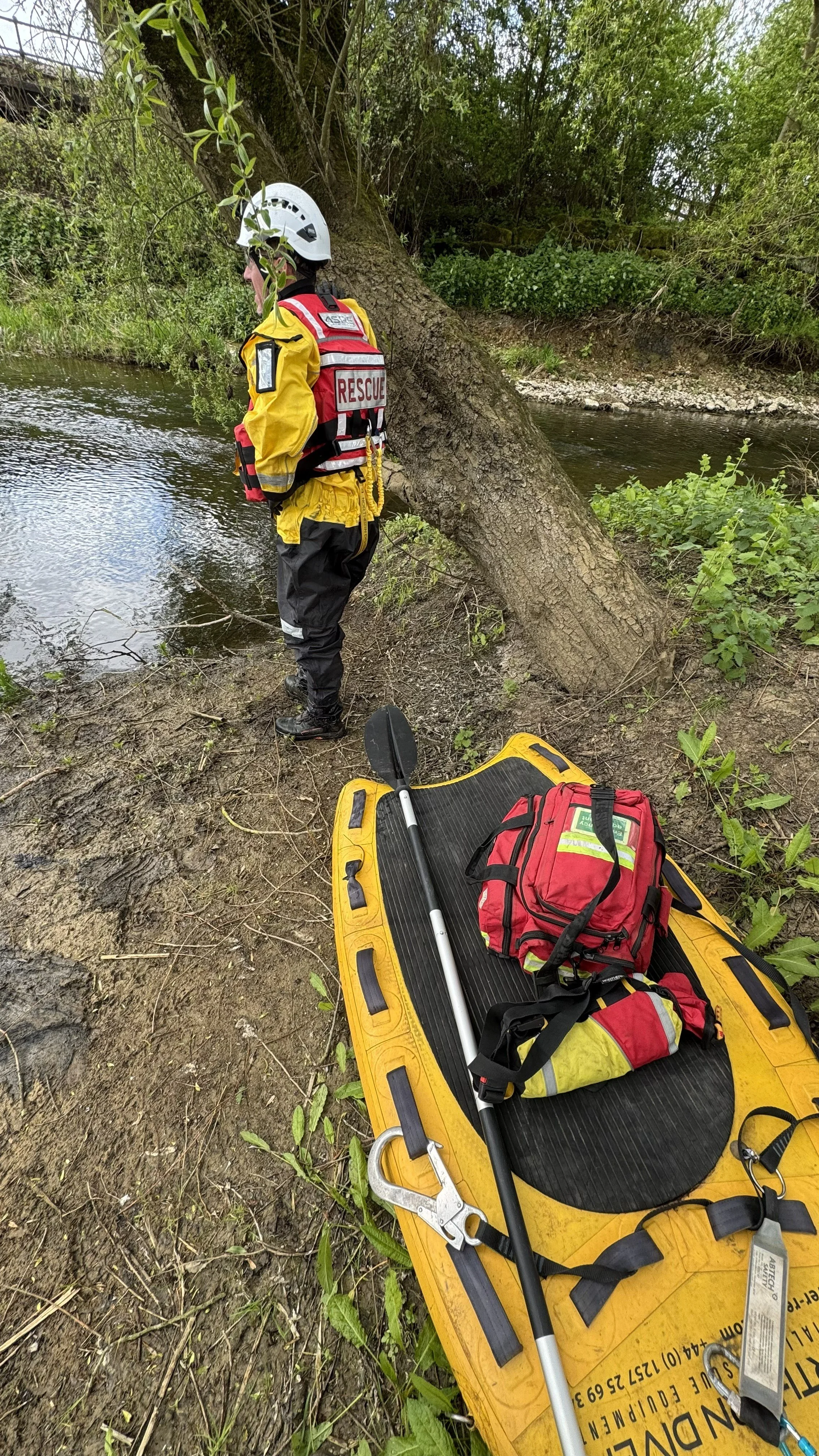 A rescue worker stands on the bank of a river next to a fallen tree. Rescue equipment, including a yellow rescue board, red bag, and metal hook, is laid on the board.