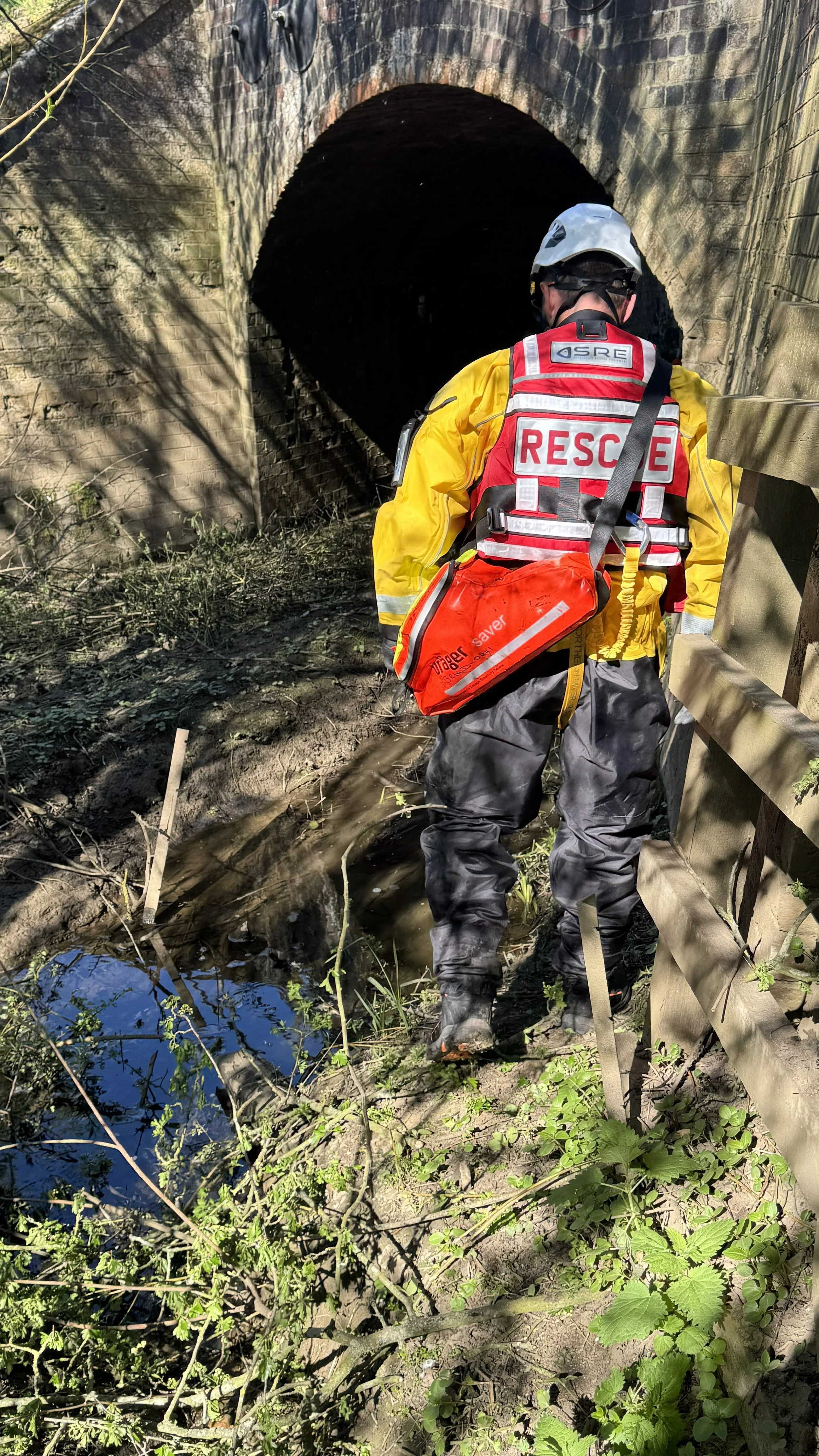 A rescue worker dressed in protective gear walking through a muddy area under a bridge, near a small stream or puddle.