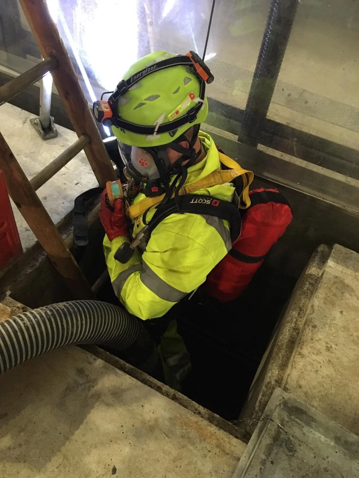 Emergency responder wearing a yellow high-visibility jacket and helmet, kneeling at an open manhole in a concrete floor, with ventilation hose nearby.