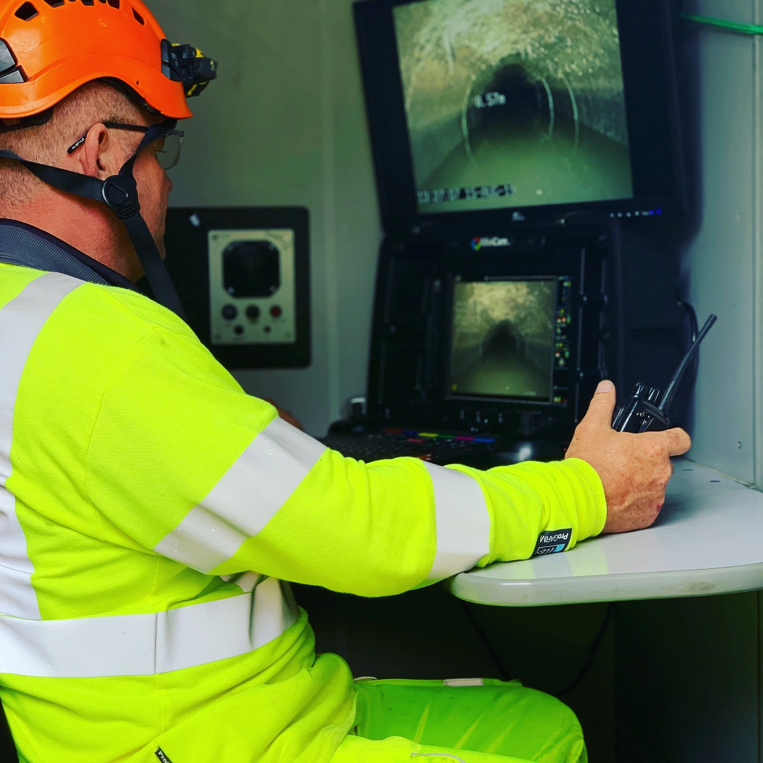 A worker in a high-visibility yellow jacket and orange helmet operating a remote control in front of computer monitors showing a tunnel view.