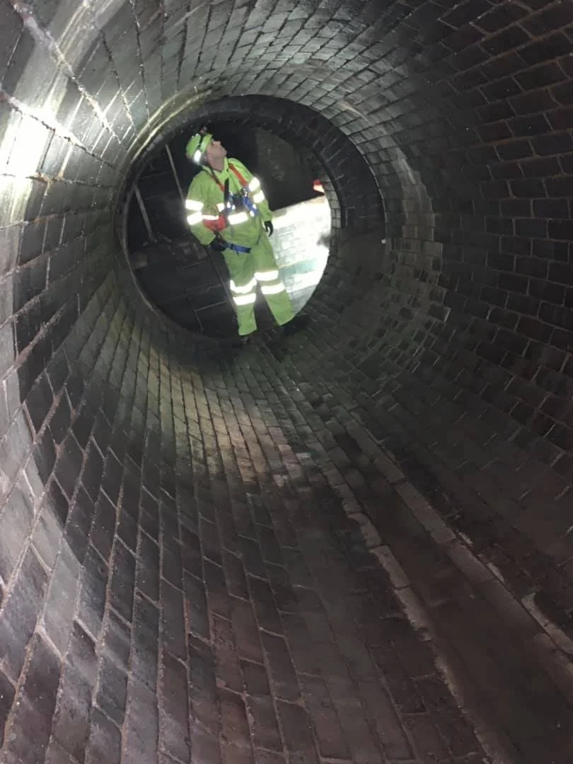 A worker in high-visibility safety gear, including a helmet and reflective stripes, standing inside a long, narrow, brick-lined sewer or tunnel.